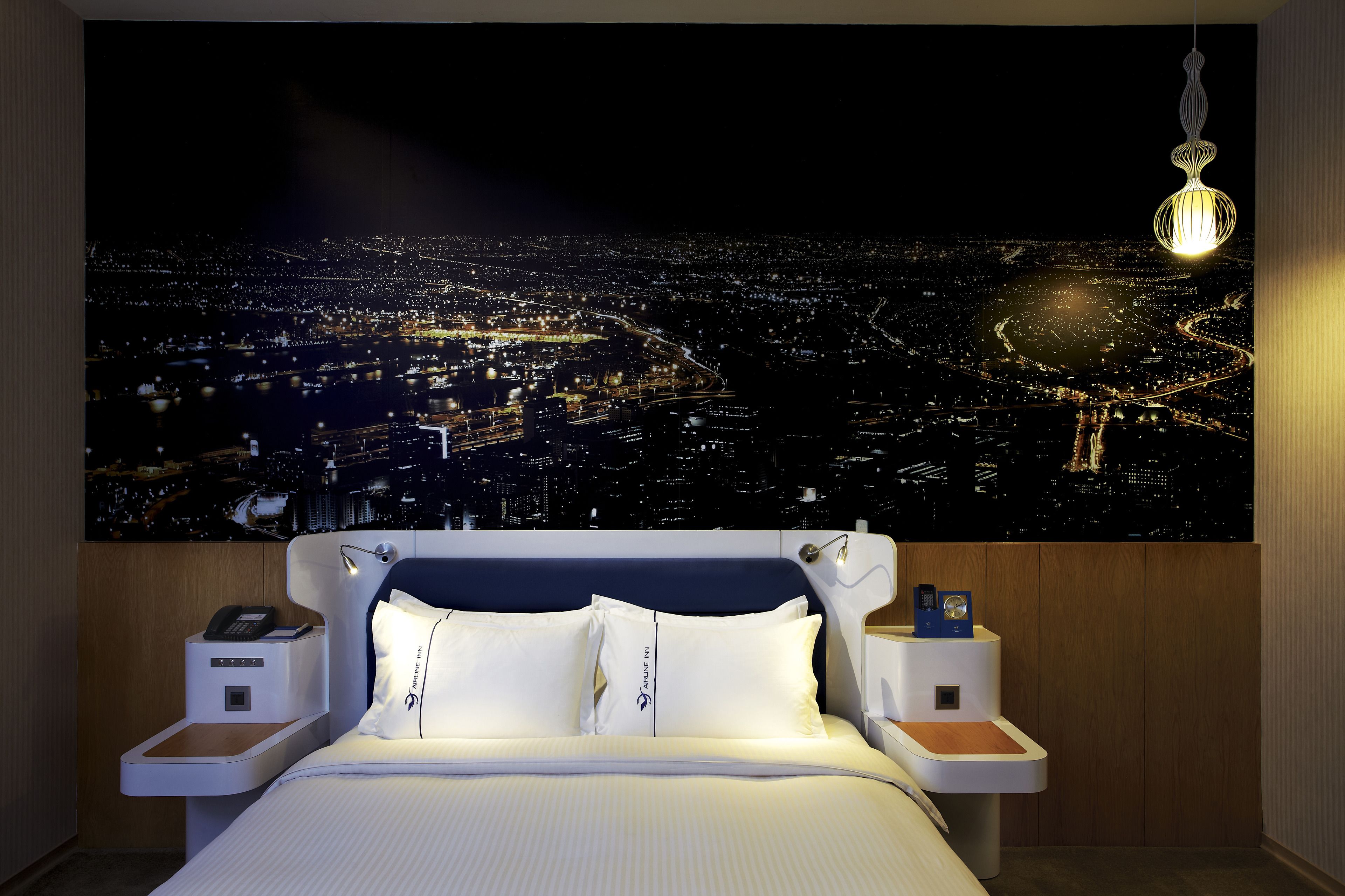 Habitación de hotel con amplia vista panorámica de la ciudad, una cama moderna, mesitas de noche con lámparas y una lámpara colgante.