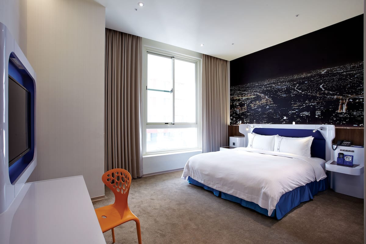 Une chambre d’hôtel avec un lit, un bureau, une chaise, une fenêtre avec des rideaux et un tableau représentant une vue sur la ville.