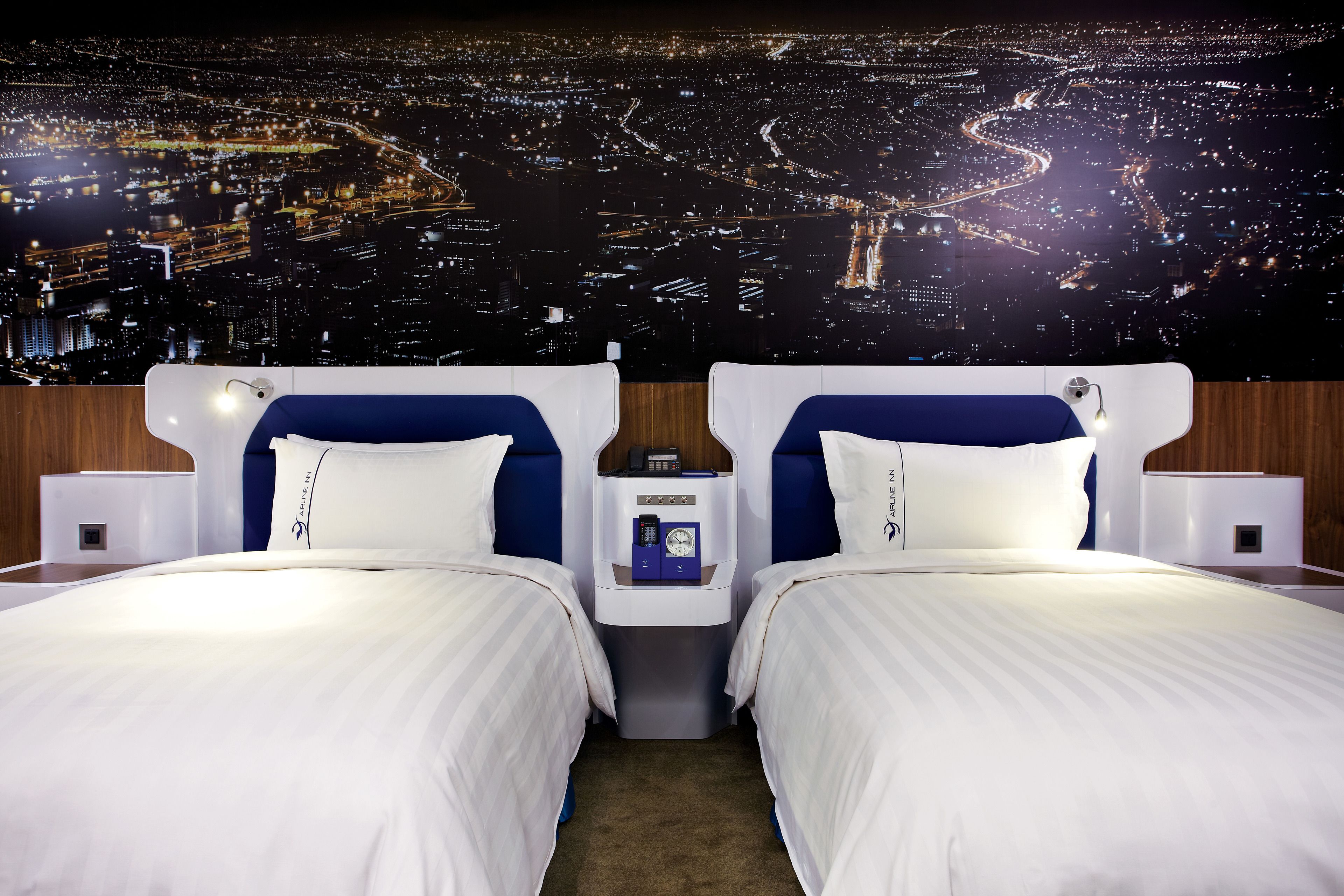A hotel room with two beds, a nightstand with a clock, and a cityscape view.