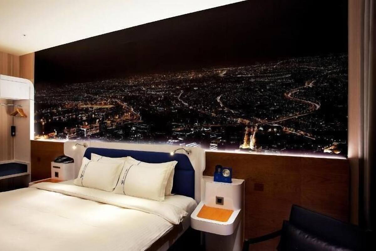 Une chambre d’hôtel comprenant un lit, des tables de chevet, une petite table, une chaise et une grande fenêtre donnant sur le paysage urbain.