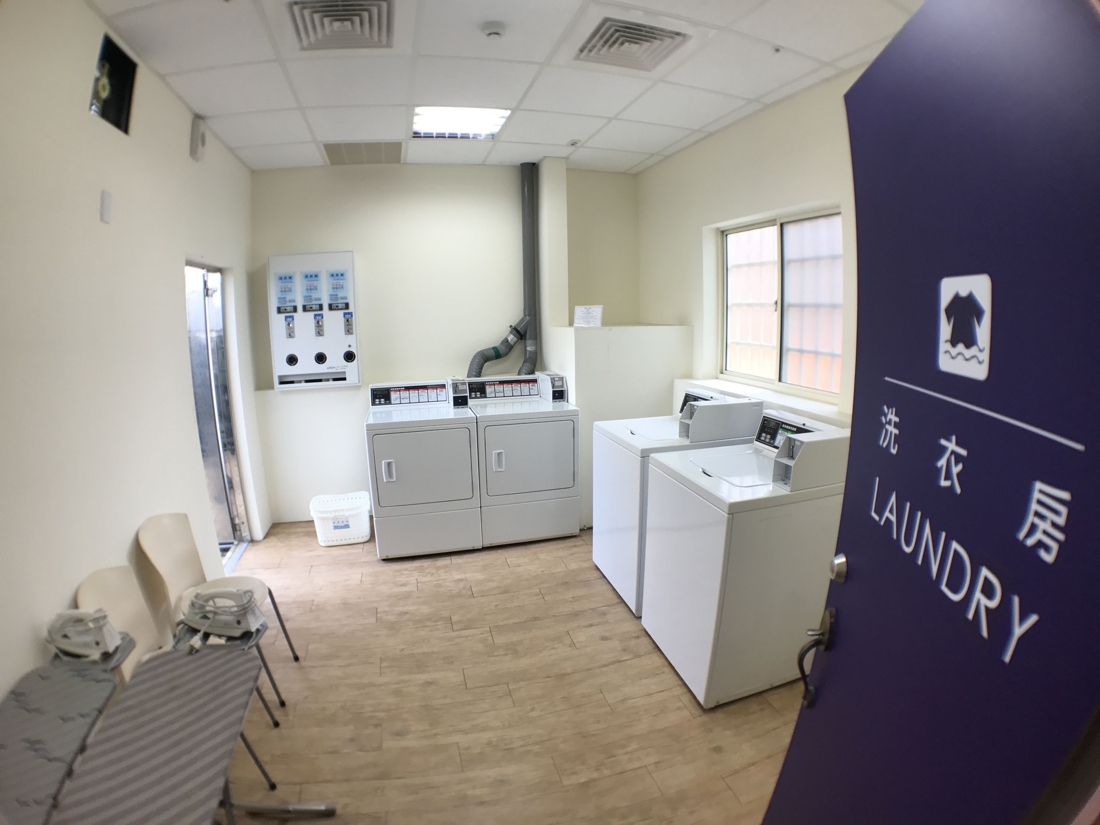 A laundry area with a washing machine, dryer, and ironing station. There is a sign indicating the laundry room, and a window with blinds.