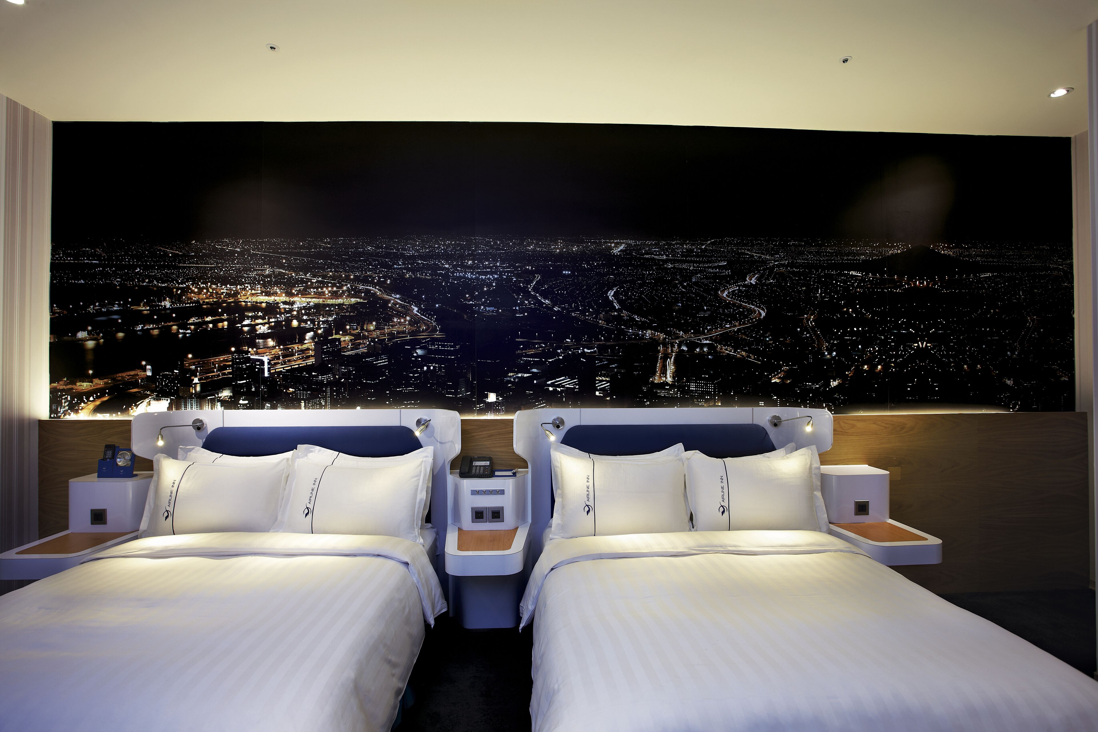 A hotel room with two beds, a nightstand, and a large cityscape view on the wall.