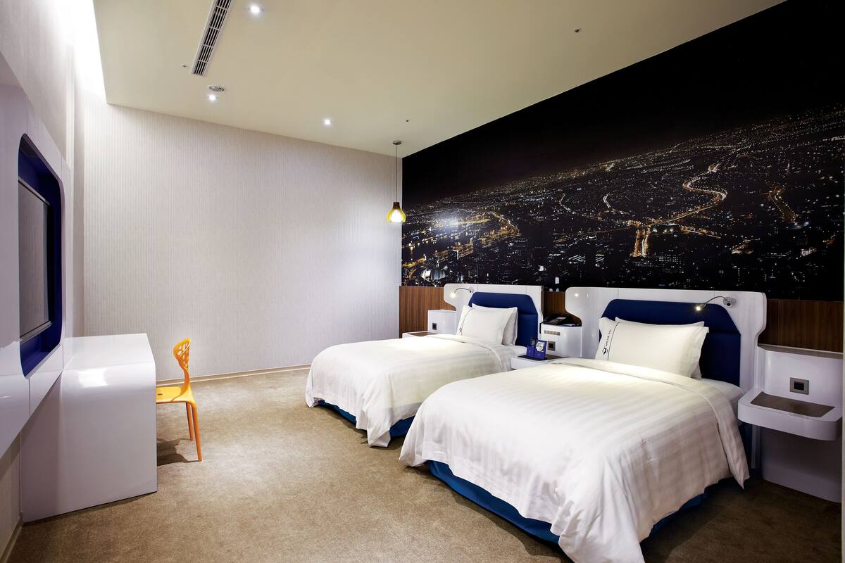 Une chambre d’hôtel moderne avec deux lits, une table de chevet, une chaise et une fresque représentant un paysage urbain sur le mur.