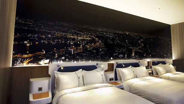 Ein modernes Hotelzimmer mit weitreichendem Stadtblick, zwei Betten und einem Nachttisch.