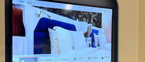 A television screen displaying a hotel room with a bed, pillows, and a nightstand.