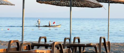 Sillas de playa y sombrillas de paja en una playa arenosa, con vista al mar y un barco al fondo.