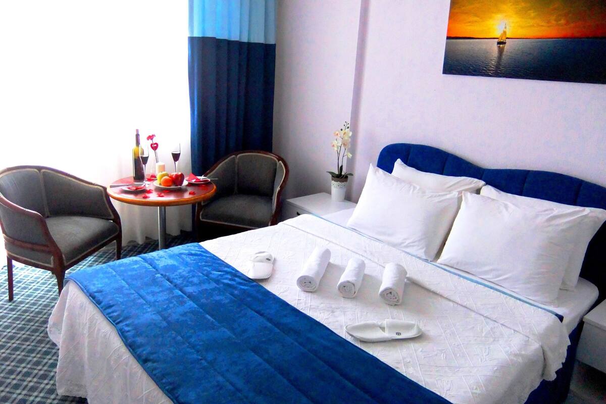 Une chambre d’hôtel avec un lit, deux chaises, une petite table sur laquelle se trouvent une bouteille et des fruits, et un tableau représentant un coucher de soleil sur l’eau.