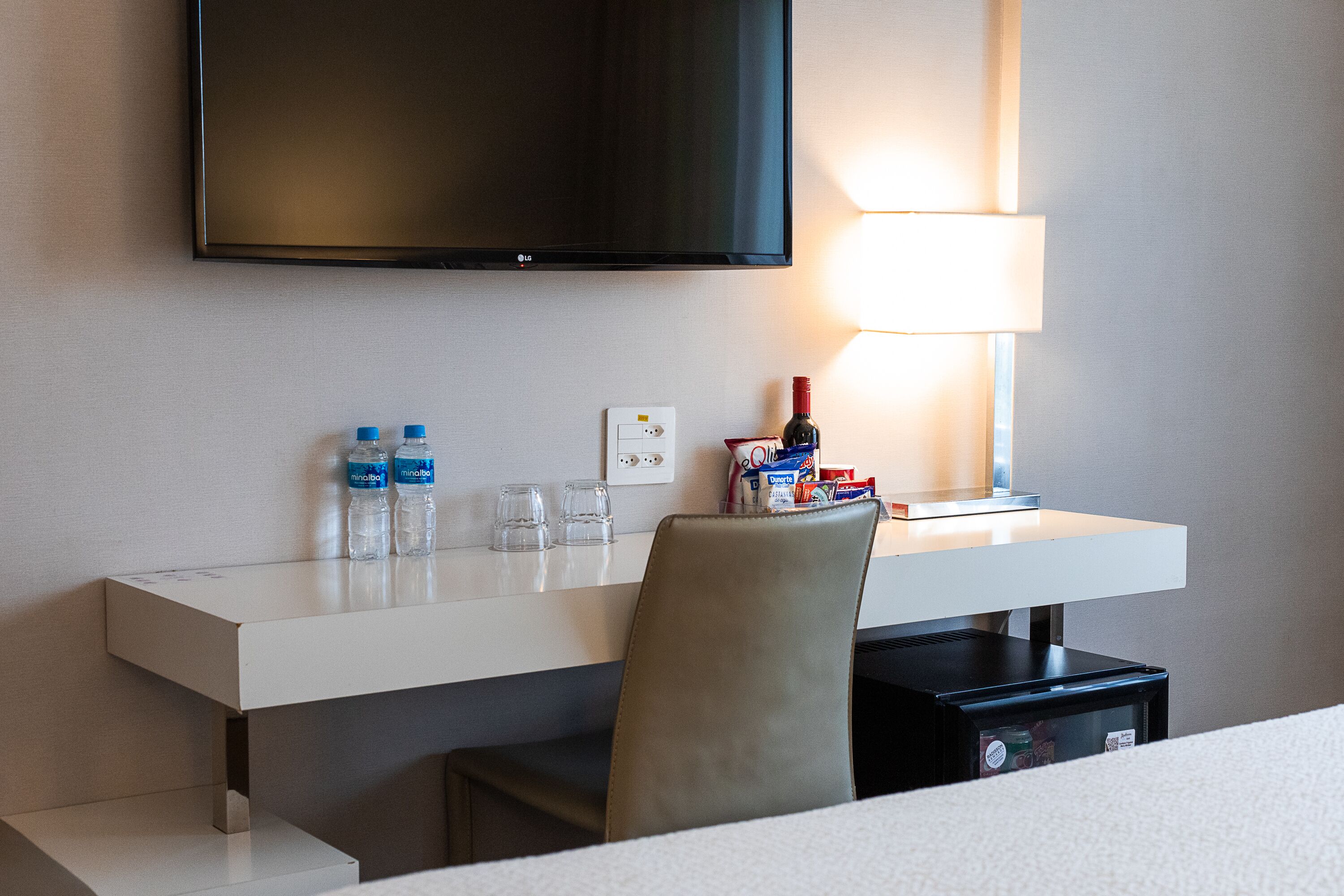Habitación de hotel con televisor de pantalla plana montado en la pared, escritorio con lámpara, una silla y un minibar.