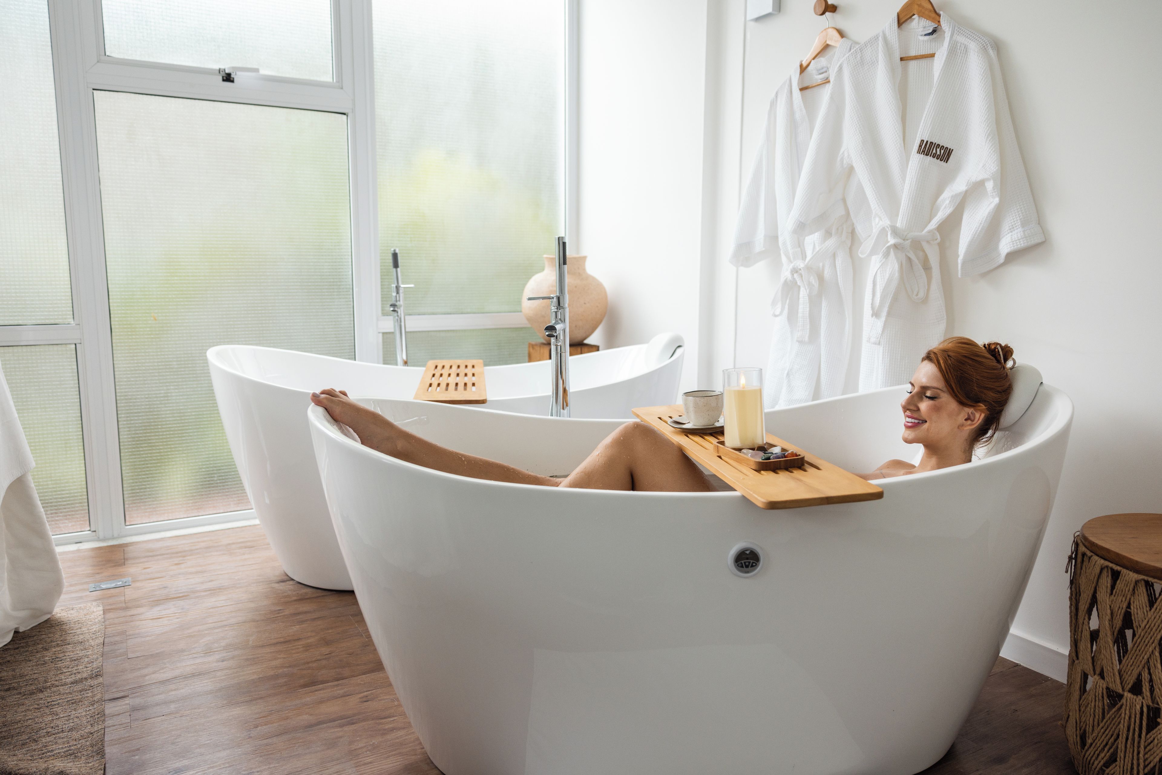 Un baño moderno con una bañera independiente, una bandeja de madera con una vela y un ventanal grande.