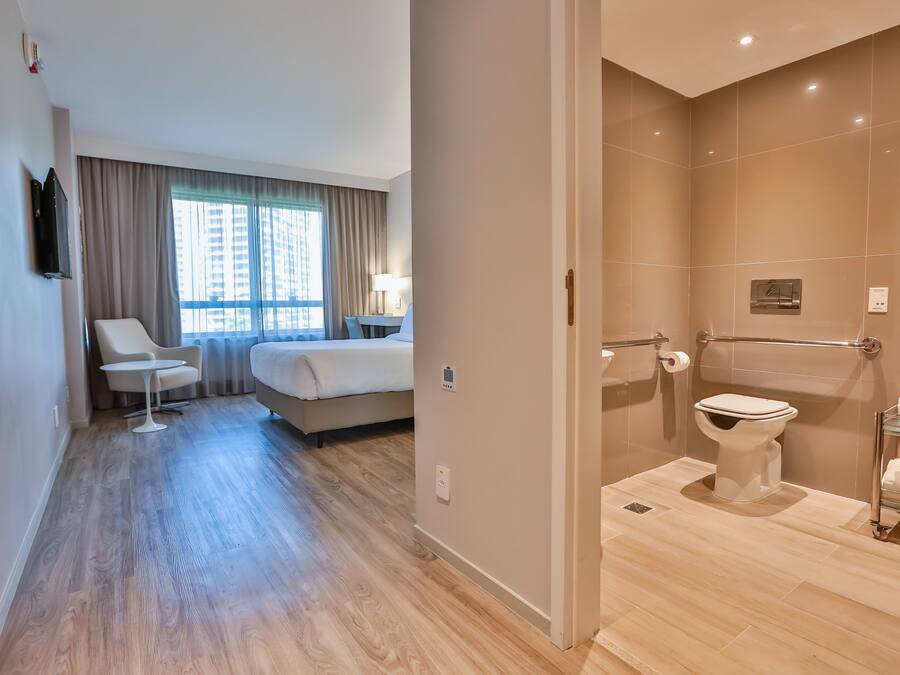 Une chambre d’hôtel comprenant un lit, une petite table, une chaise, un téléviseur et une salle de bain avec des toilettes et une douche à parois de verre.