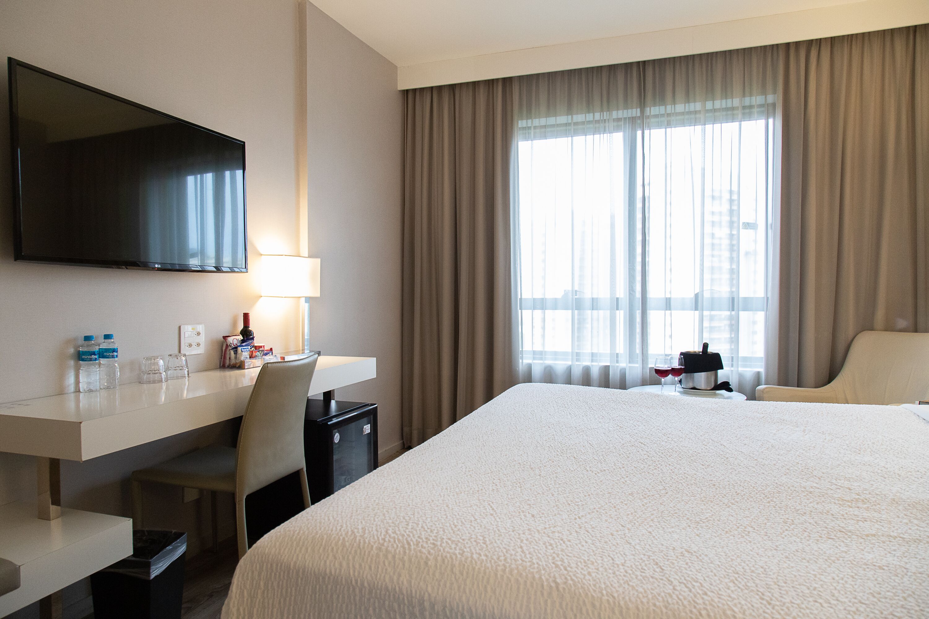 Habitación de hotel con una cama grande, un escritorio con televisión de pantalla plana, una silla, una lámpara y una ventana con cortinas.