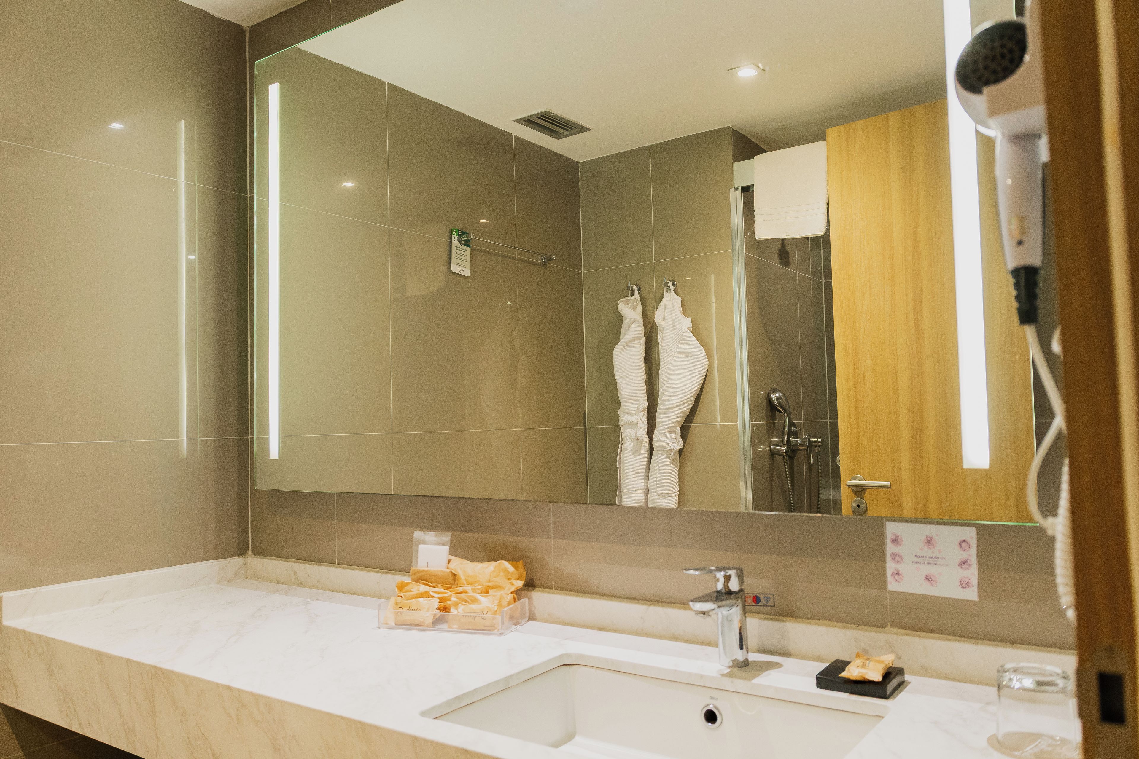 A hotel bathroom with a large mirror, a sink, a shower, and a hairdryer.