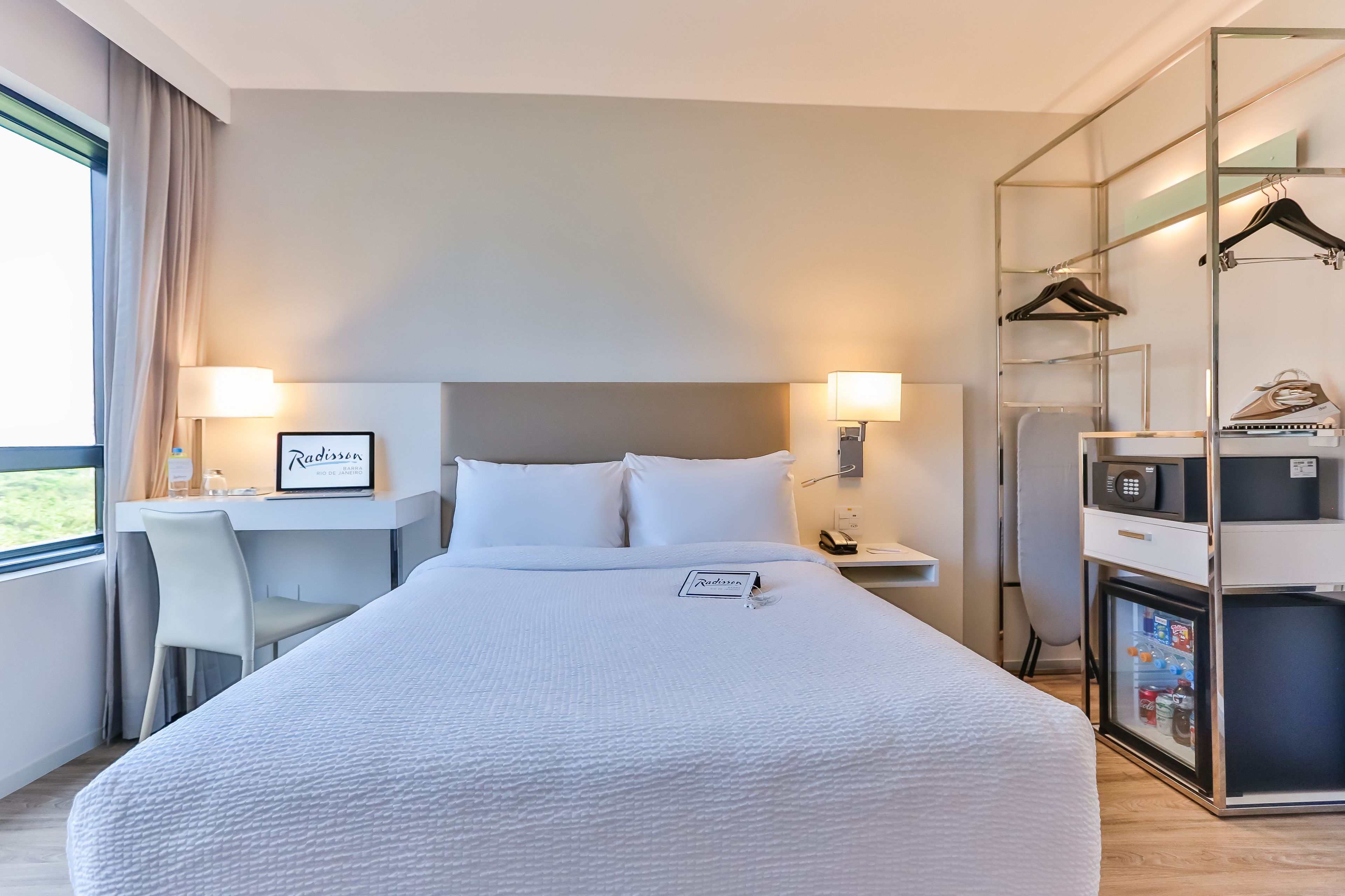 A hotel room with a large bed, a desk with a chair, a desk lamp, a nightstand with a phone, a coat rack, and a minibar.