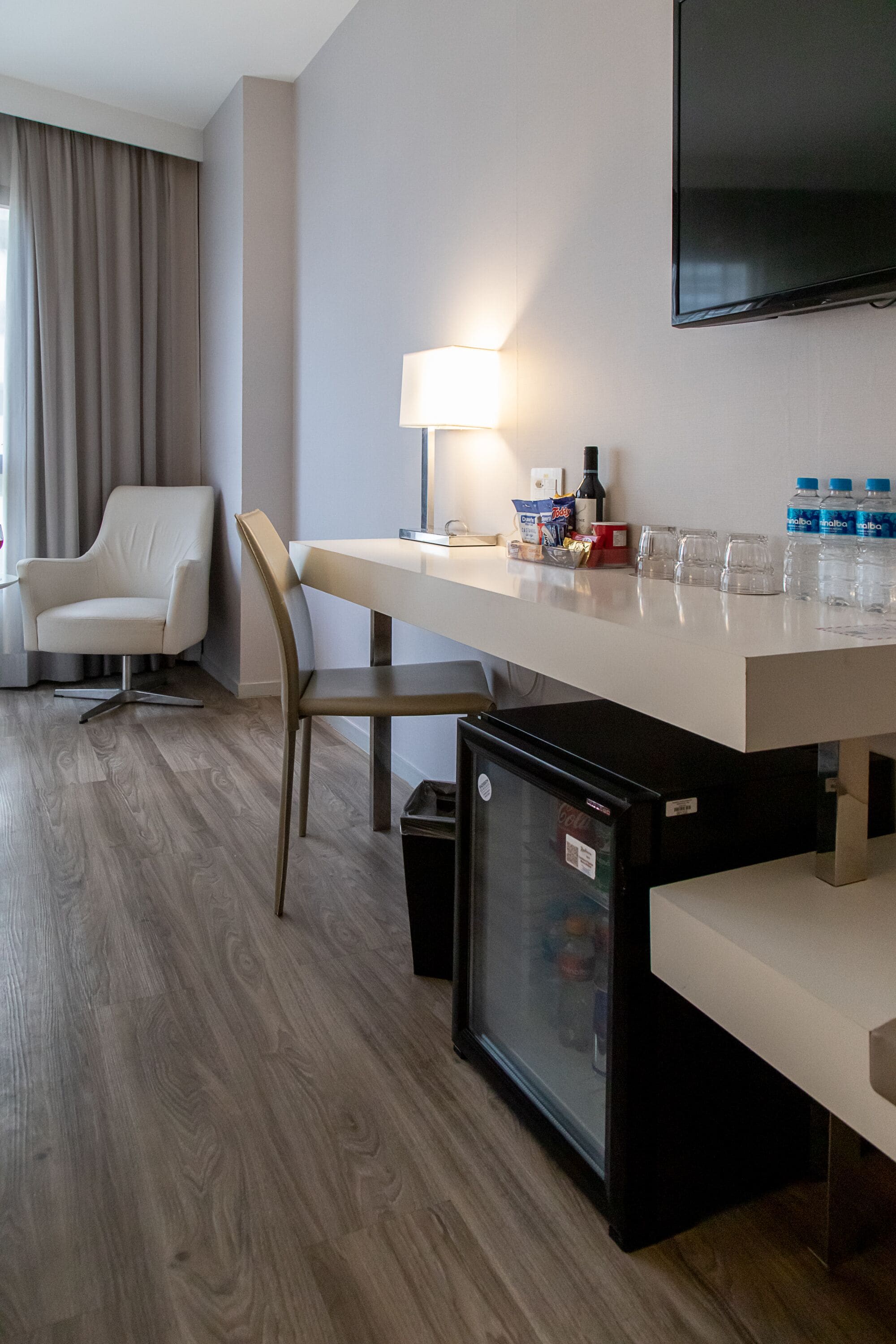 A hotel room with a wooden floor, a white desk with a lamp, a TV mounted on the wall, a mini fridge, a chair, and a window with curtains.