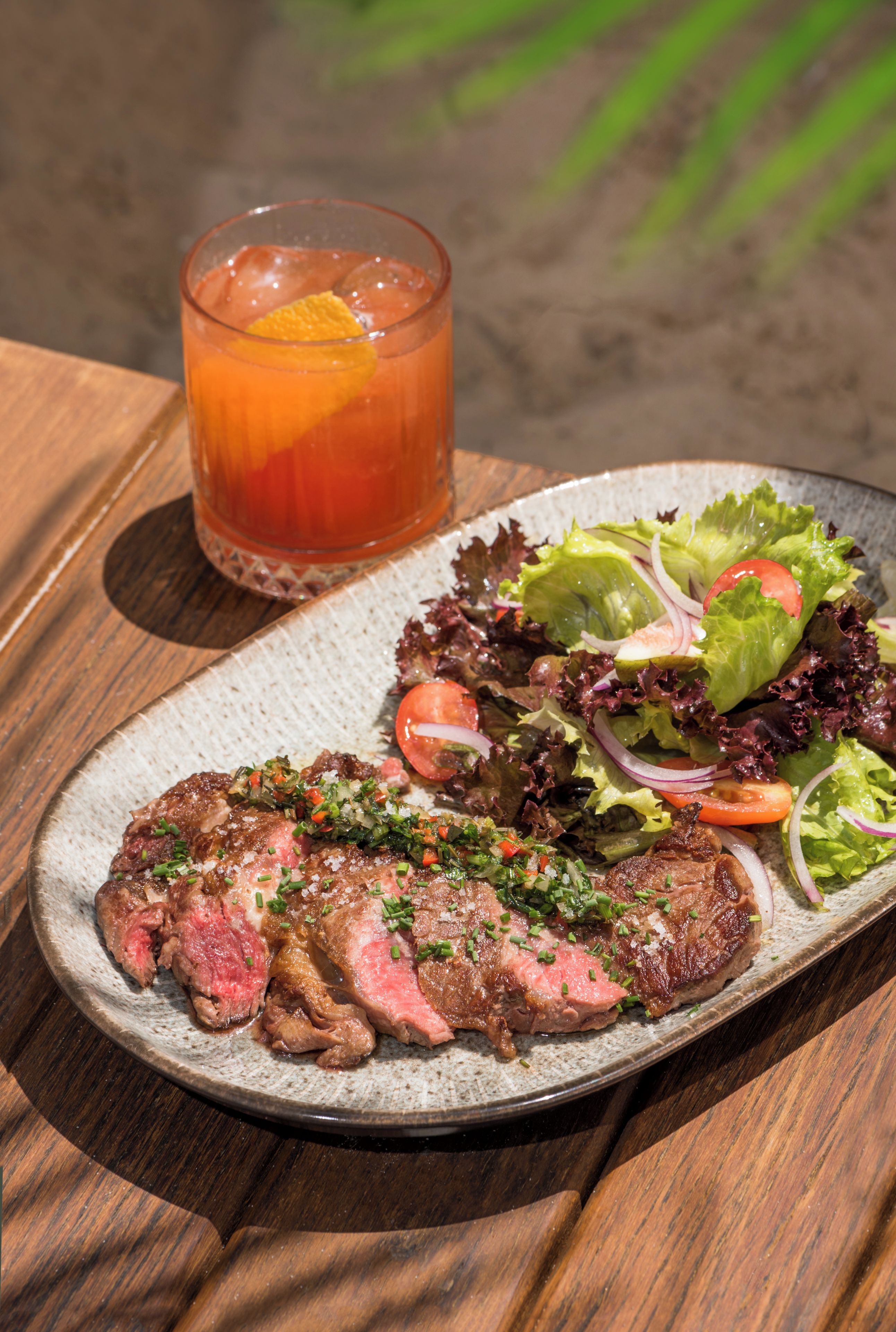 A plate with steak and salad, and a glass of beverage on a wooden table.