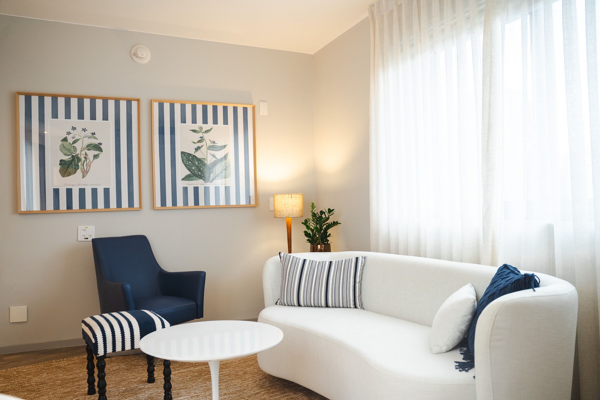 Un salon avec un canapé blanc, un fauteuil à rayures et une petite table ronde. Deux reproductions botaniques encadrées ornent le mur, et une lampe est posée sur une table d’appoint.