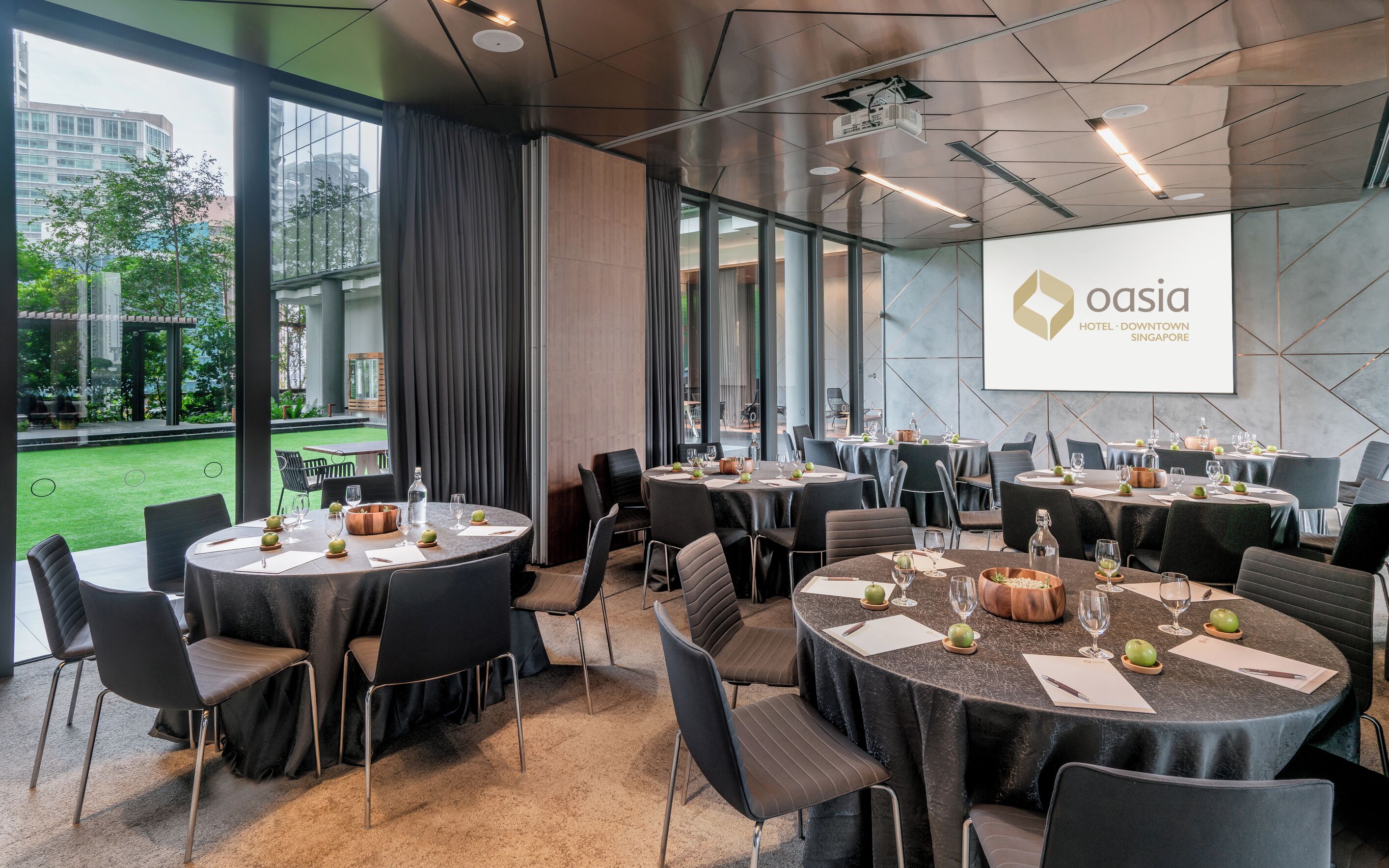 Een vergaderruimte met meerdere tafels gedekt voor een maaltijd, grote ramen en een presentatiescherm waarop de naam van het hotel staat.