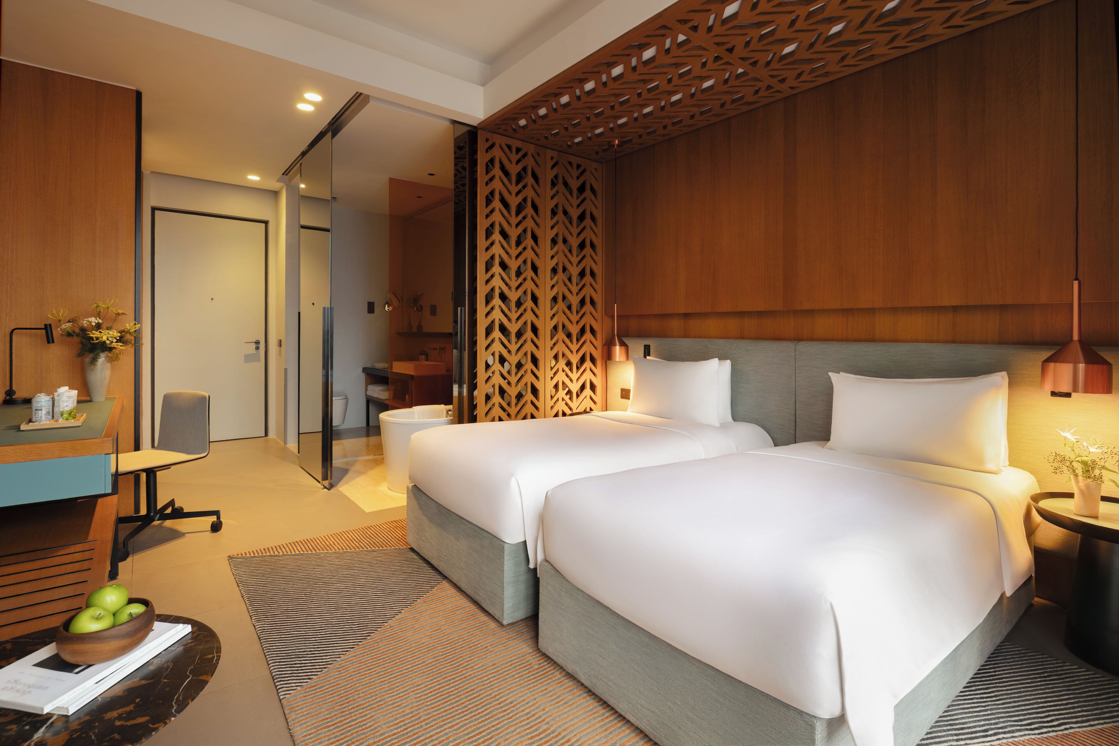 Habitación de hotel con dos camas, un escritorio con silla, una mesita con un florero y un panel decorativo en la pared.