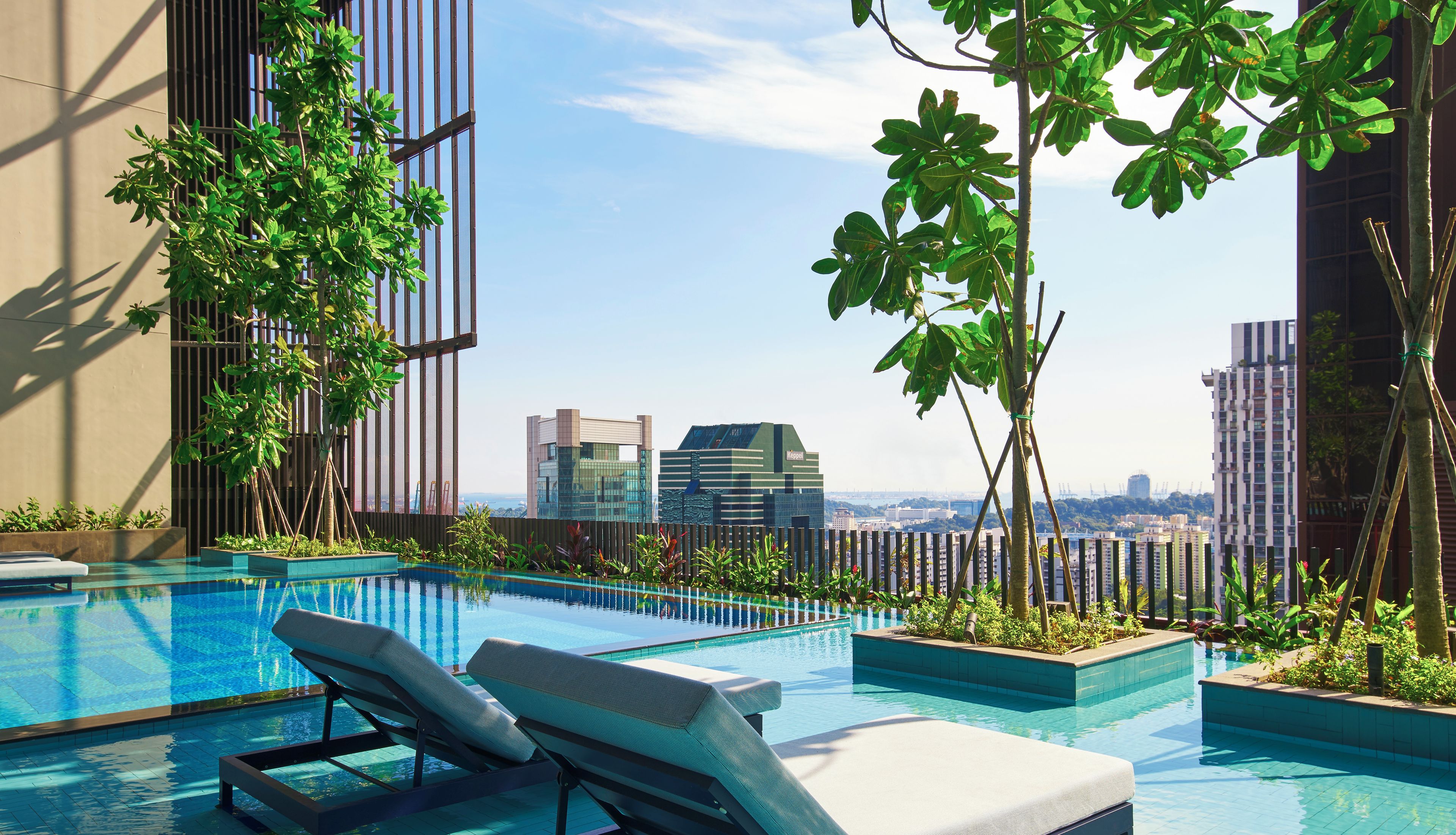 A rooftop infinity pool with lounge chairs, surrounded by greenery and overlooking a cityscape.