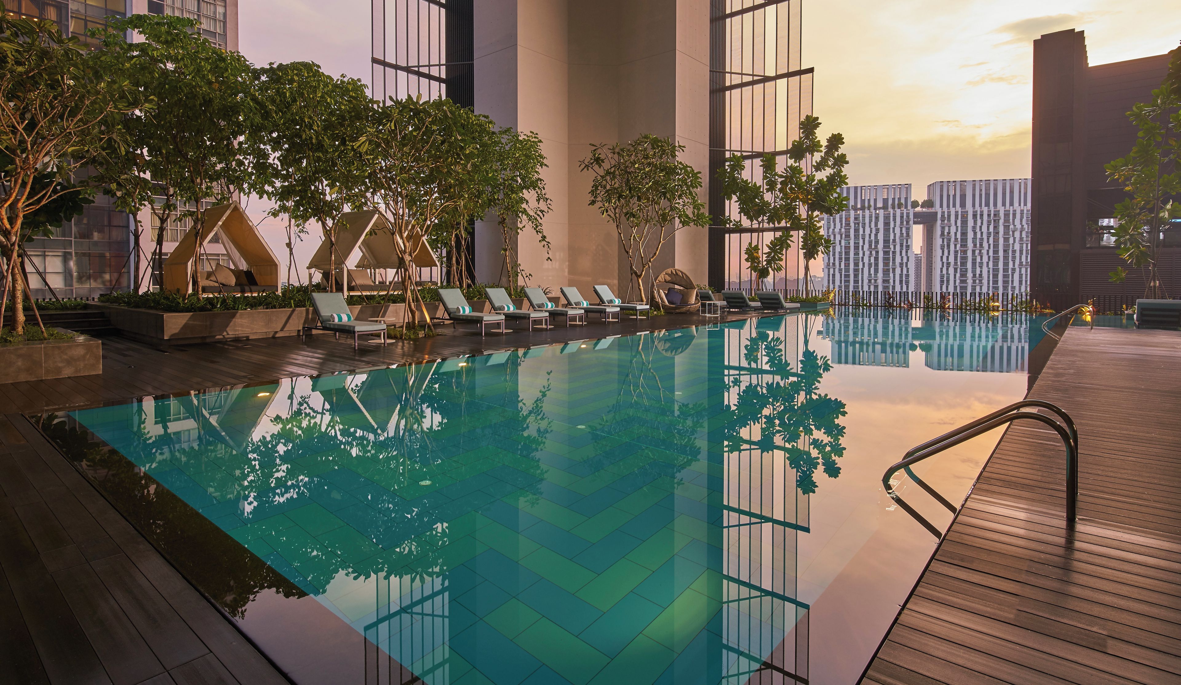 An infinity pool with lounge chairs, surrounded by trees and modern buildings.