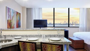 A modern hotel room with a granite countertop, two sinks, a flat-screen TV, a bed with a view of the city skyline, and abstract art on the walls. - Residence Inn by Marriott Rio de Janeiro Barra da Tijuca (Rio de Janeiro)