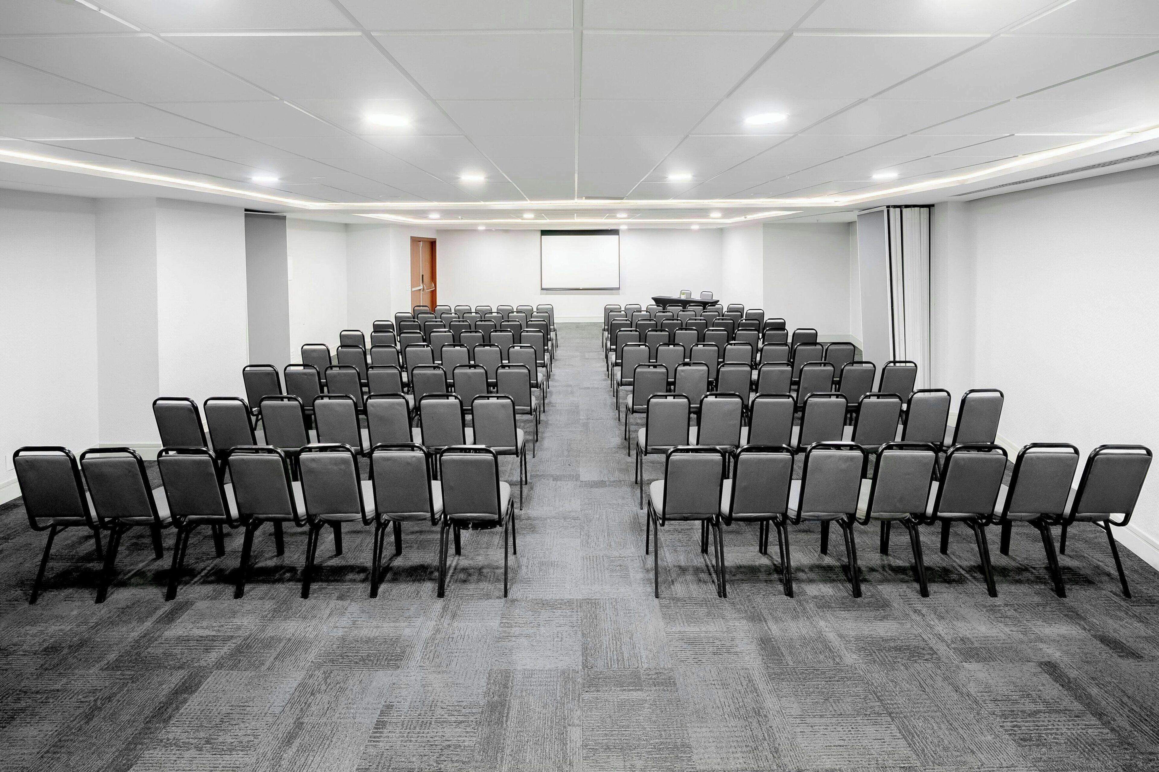 A conference room with rows of chairs, a projector screen, and a carpeted floor.