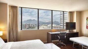 A hotel room with two beds, a desk with a chair, a TV, and a large window offering a city view.