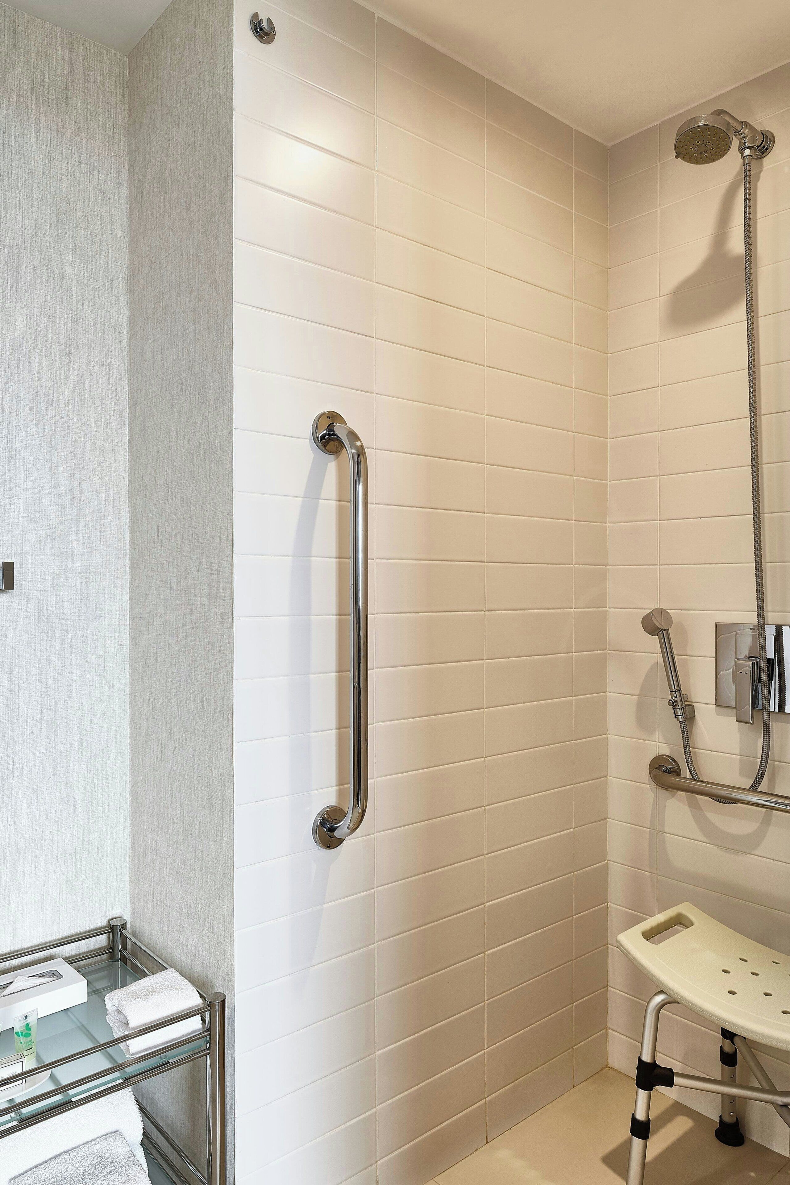 Une salle de bain moderne avec des murs recouverts de carreaux blancs, une douche à parois de verre, un pommeau de douche à main et une étagère en verre où sont posées des serviettes.