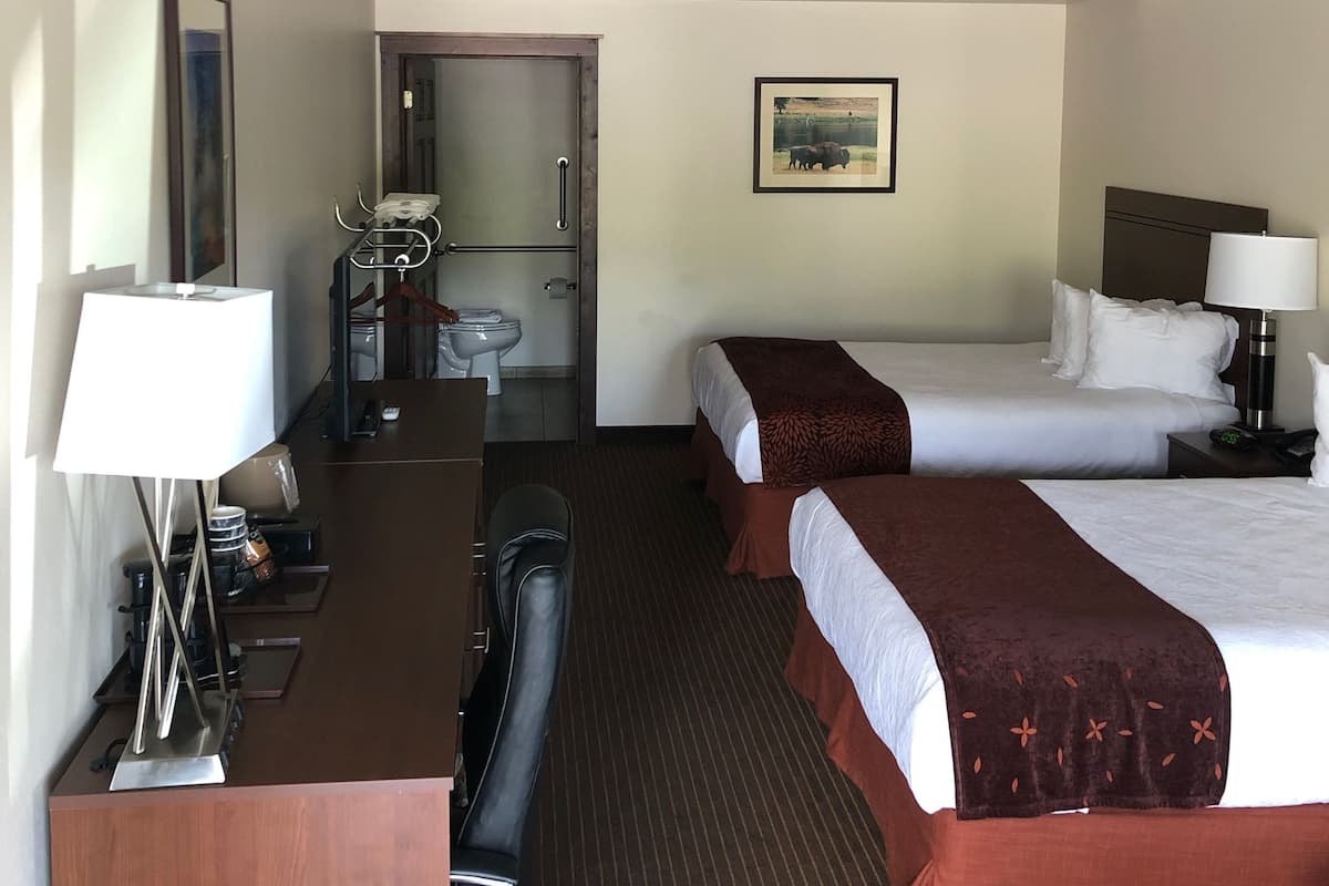 Habitación de hotel con dos camas, un escritorio con lámpara, una silla y un baño visible a través de una puerta abierta.