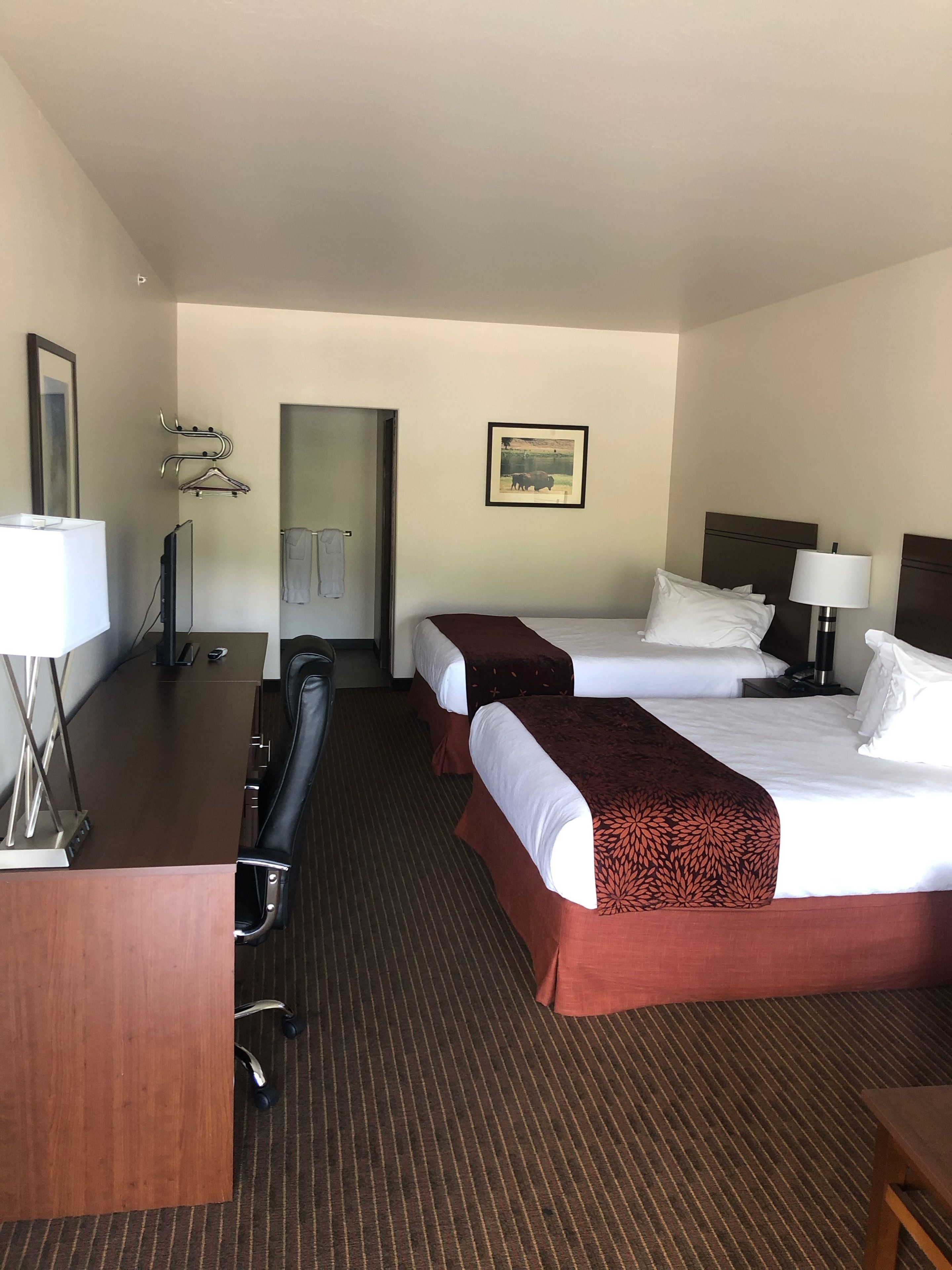 A hotel room with two beds, a desk, a chair, a lamp, and a painting on the wall.