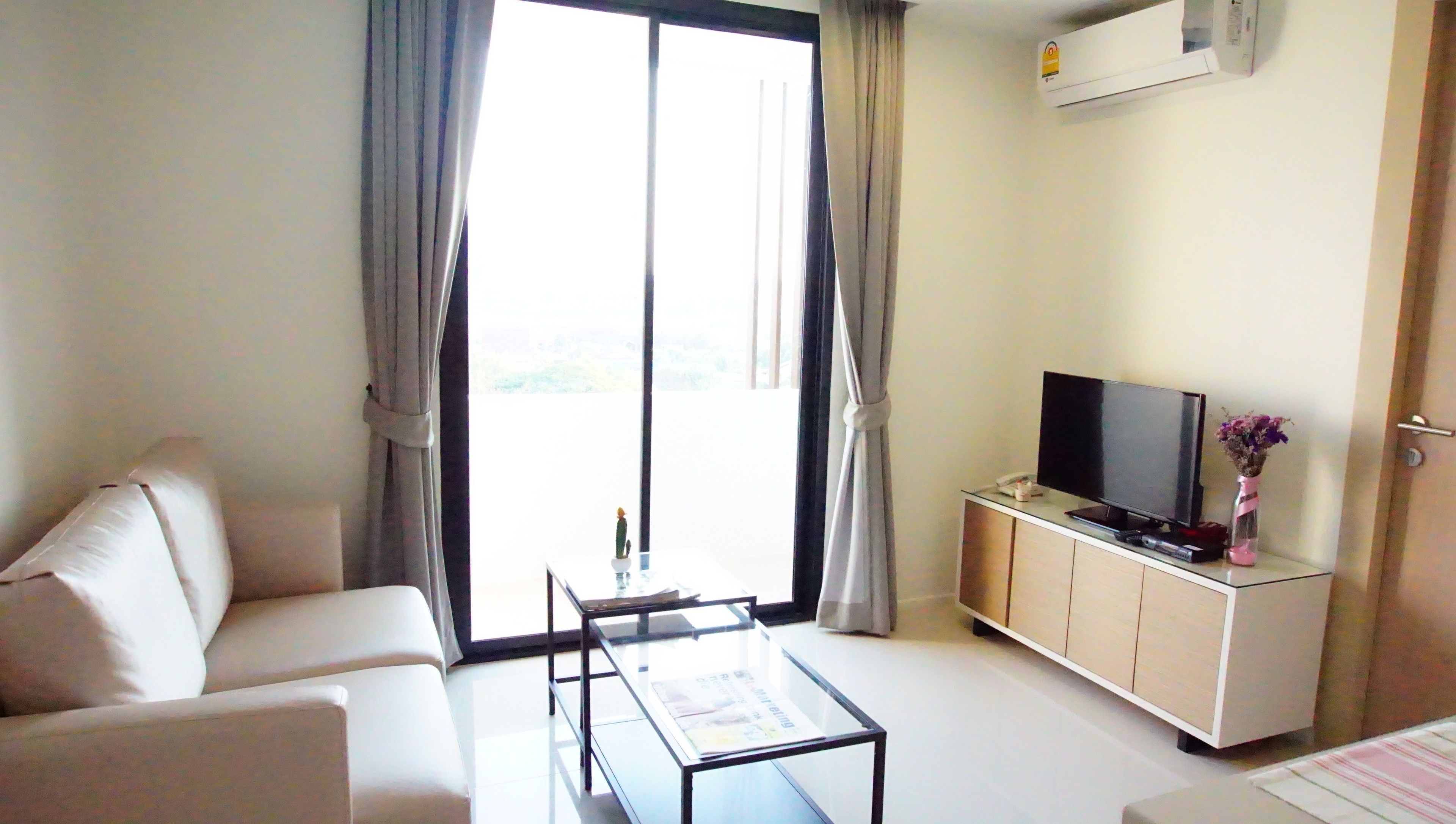 Una sala de estar moderna con un sofá, una mesa de centro de vidrio, un televisor de pantalla plana sobre un mueble de madera y un gran ventanal corredero que da a un balcón.