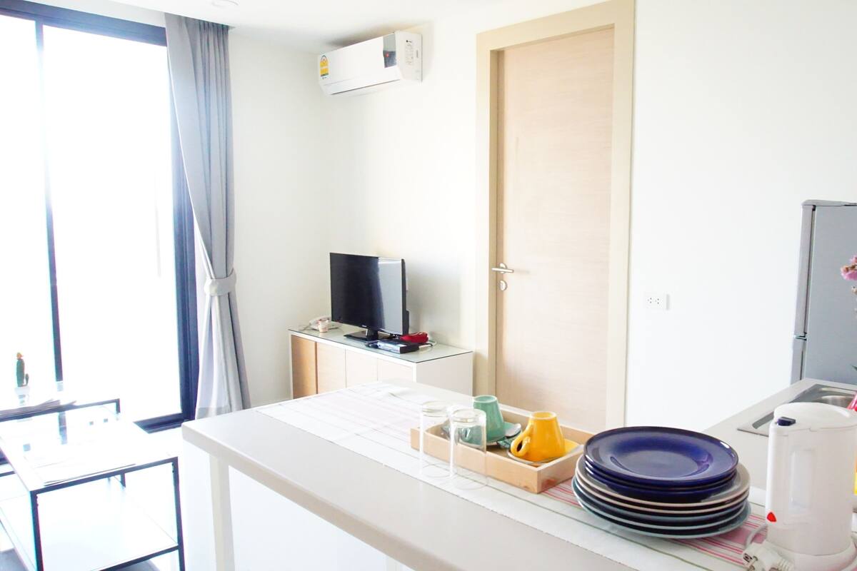 A hotel room with a bed, a TV, a desk with a computer, a window with curtains, and a vase with flowers.
