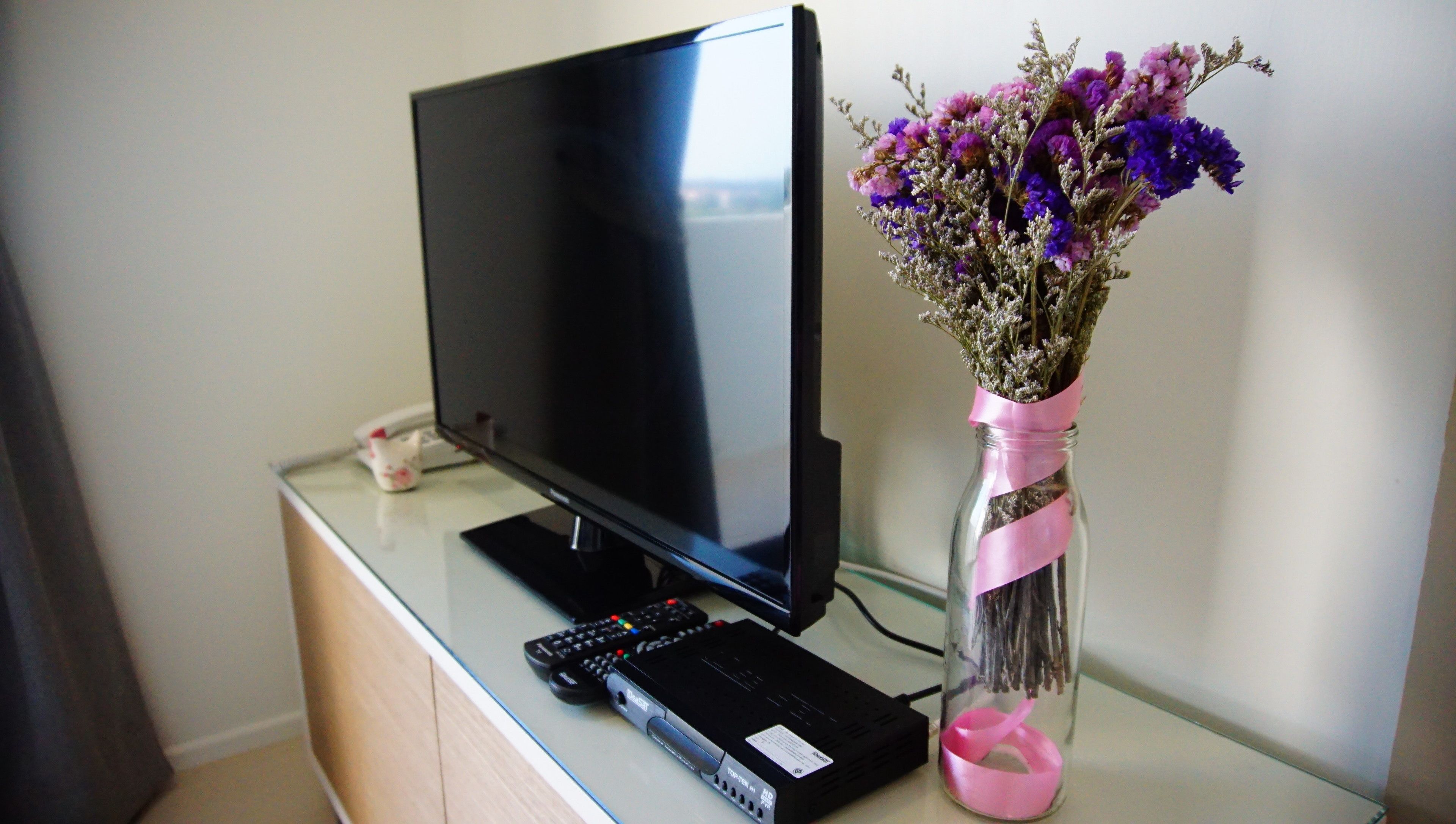 A flat-screen TV on a glass table with a remote control and a decorative vase with flowers.