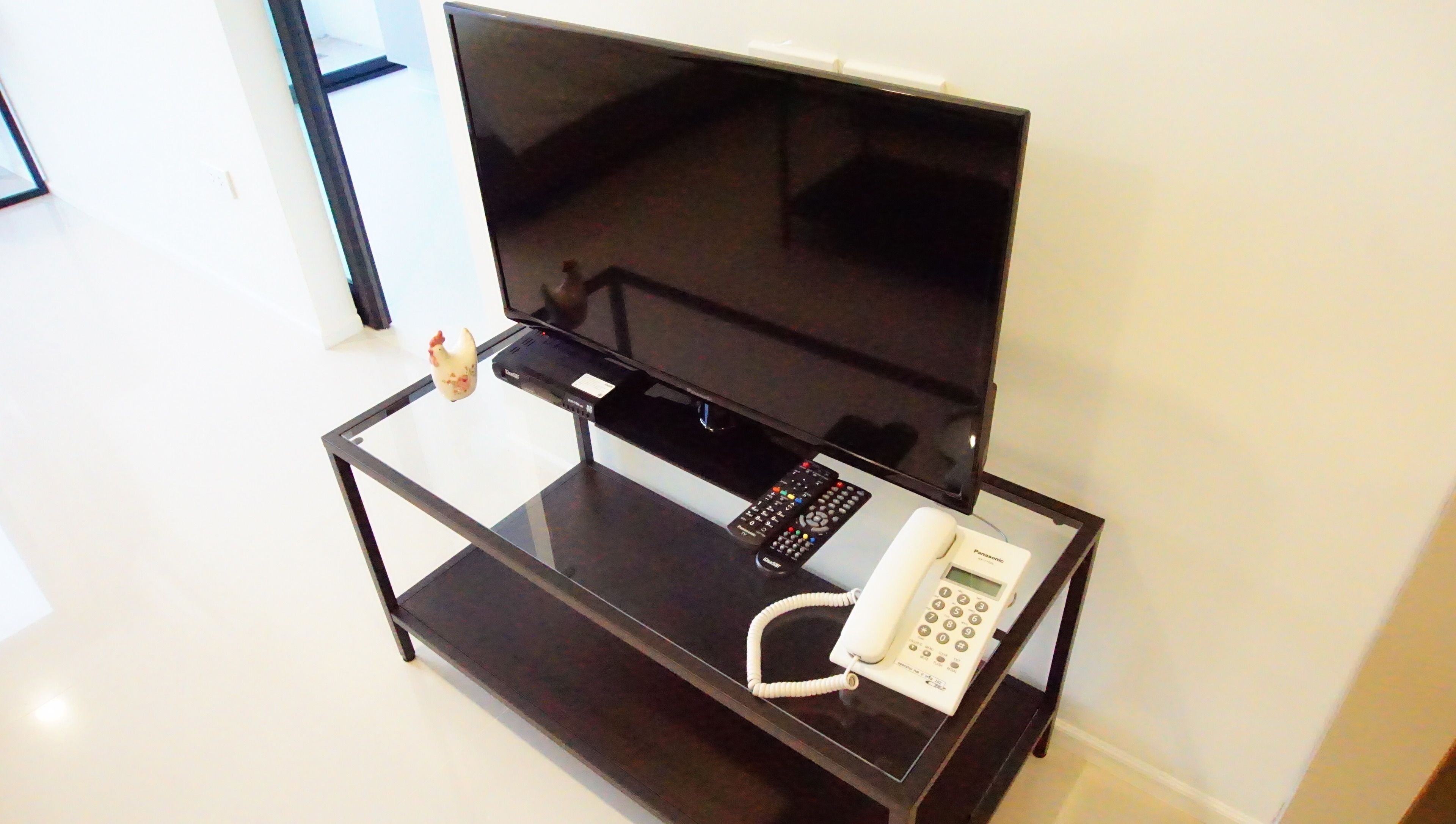 A glass-top table with a flat-screen TV, a remote control, and a white telephone.