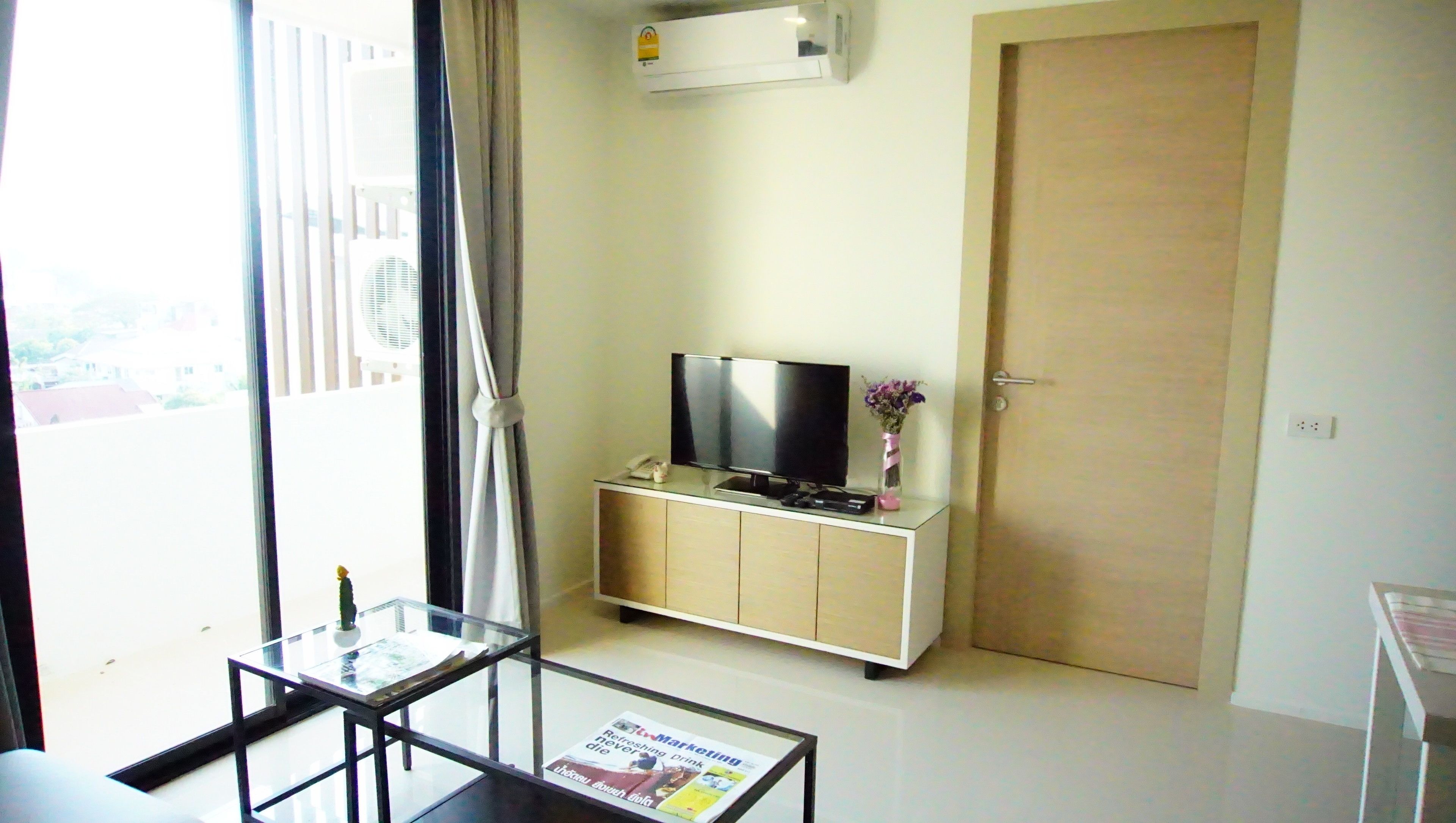 A modern hotel room with a flat-screen TV, a small cabinet, a glass table, and a door.