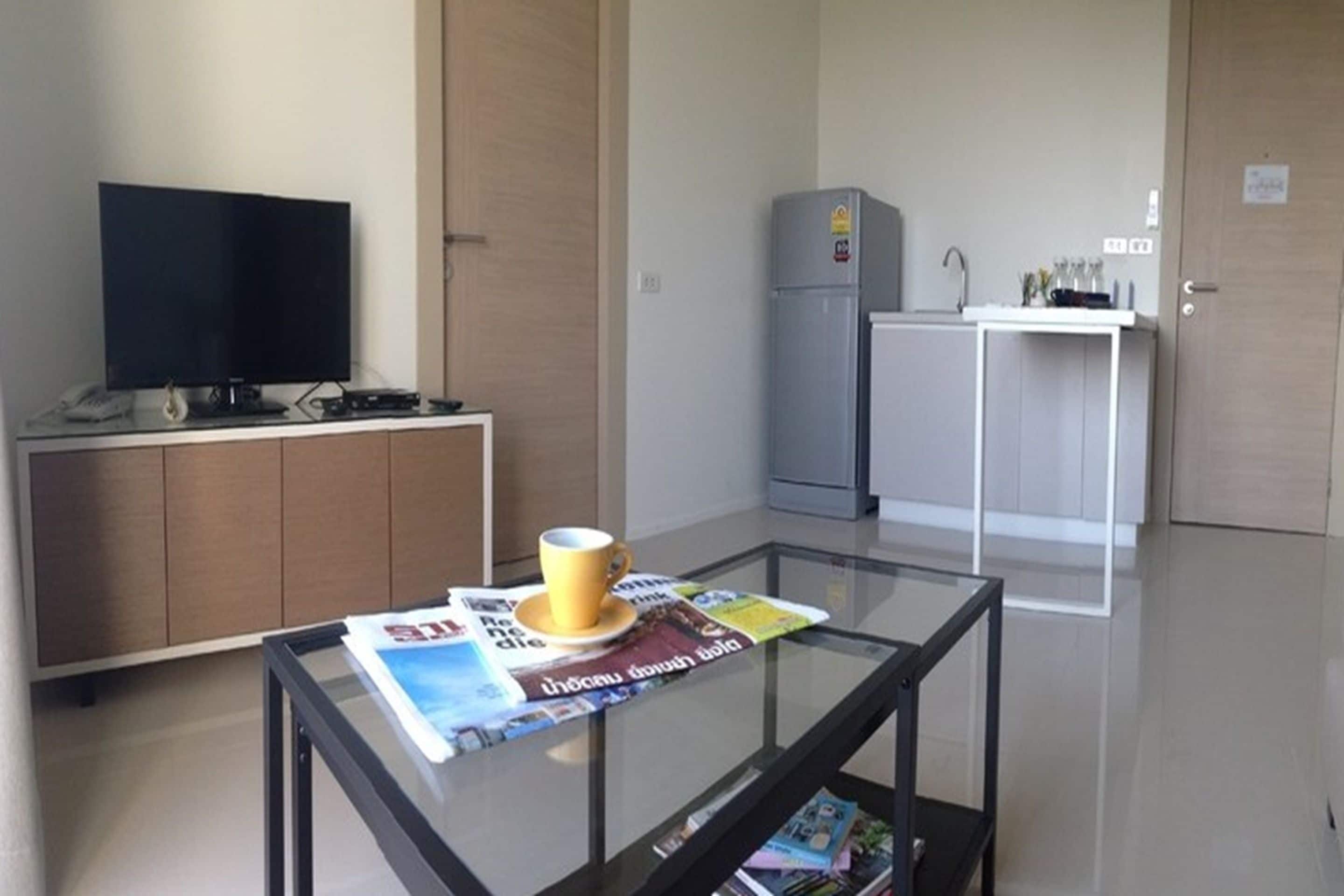 A modern living room with a glass coffee table, a flat-screen TV, a kitchen area with a refrigerator and sink, and a wooden cabinet.
