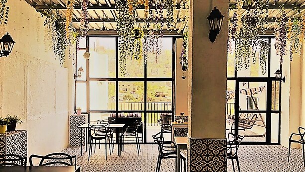 Un café con baldosas de diseño, plantas colgantes y vistas amplias a través de grandes ventanales.