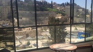 Una habitación con un ventanal grande que ofrece vistas a un paisaje urbano, incluyendo edificios y una colina. Hay dos sillas de madera y una mesita redonda con un mantel individual.