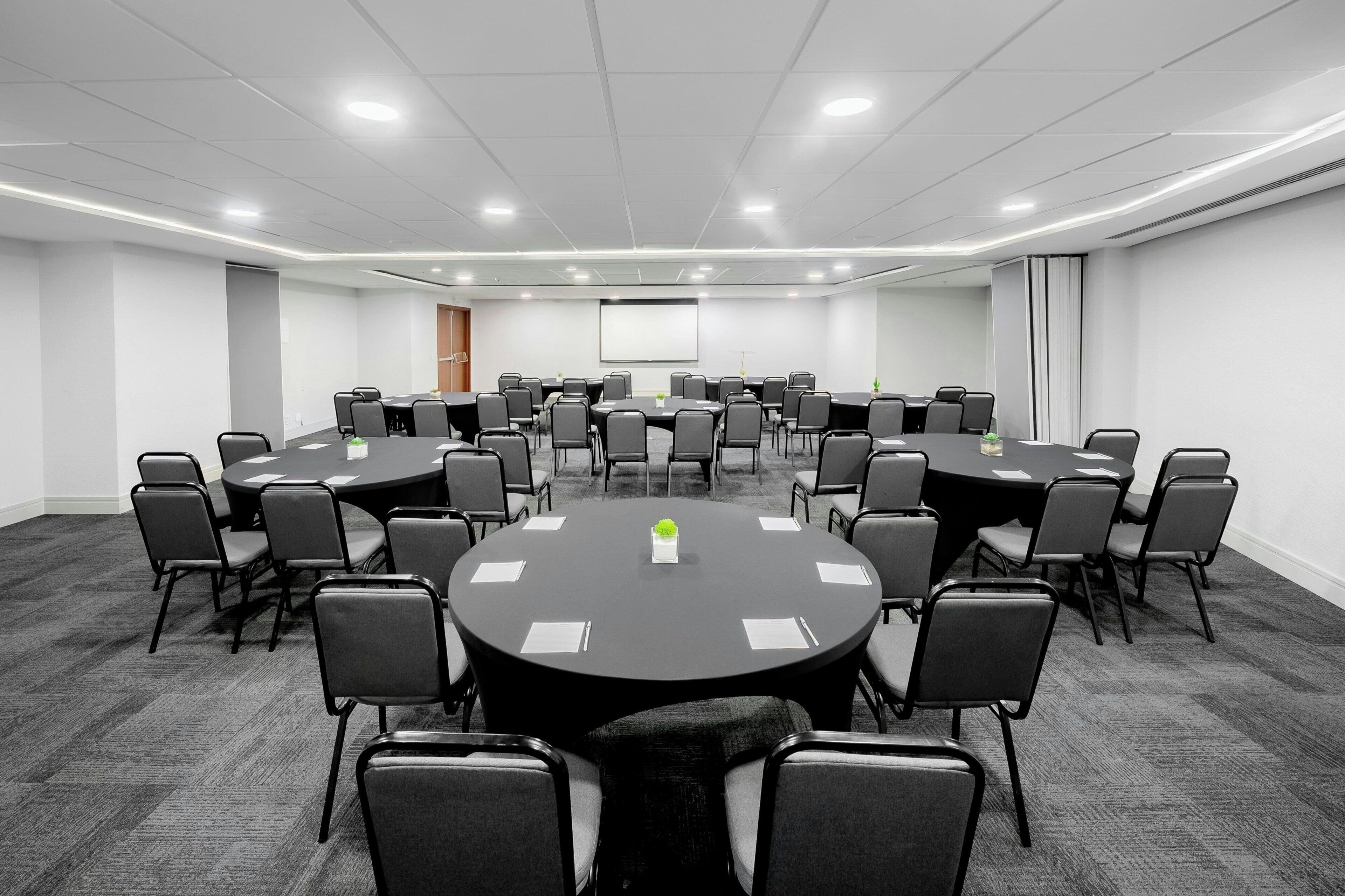 A conference room with multiple tables and chairs, a projector screen, and a carpeted floor.