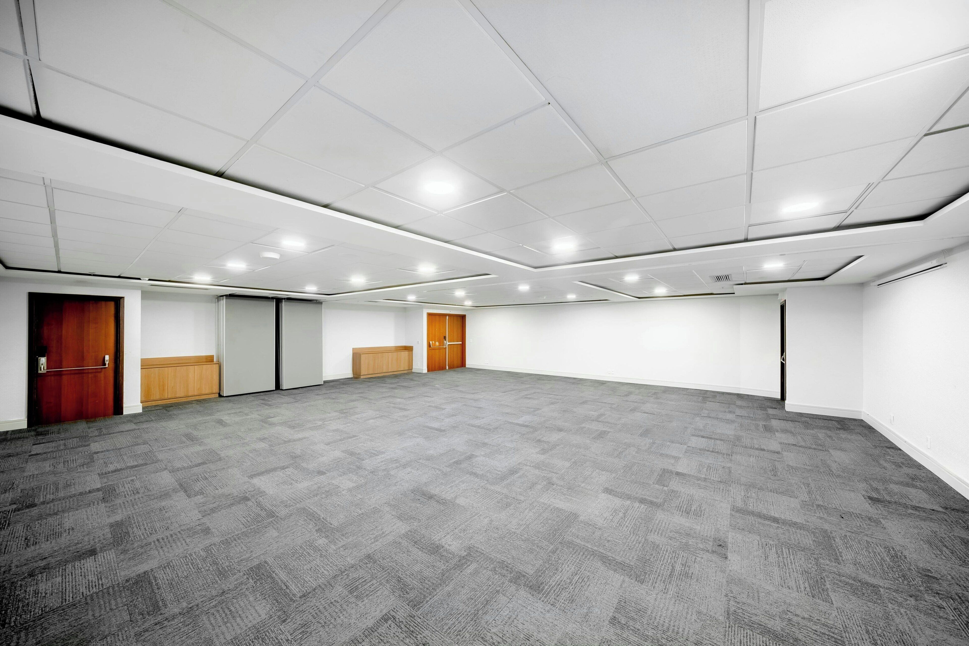 A large, empty conference room with carpeted flooring, white walls, and a grid of ceiling lights.
