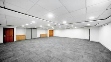 A large, empty conference room with carpeted flooring, white walls, and a grid of ceiling lights.