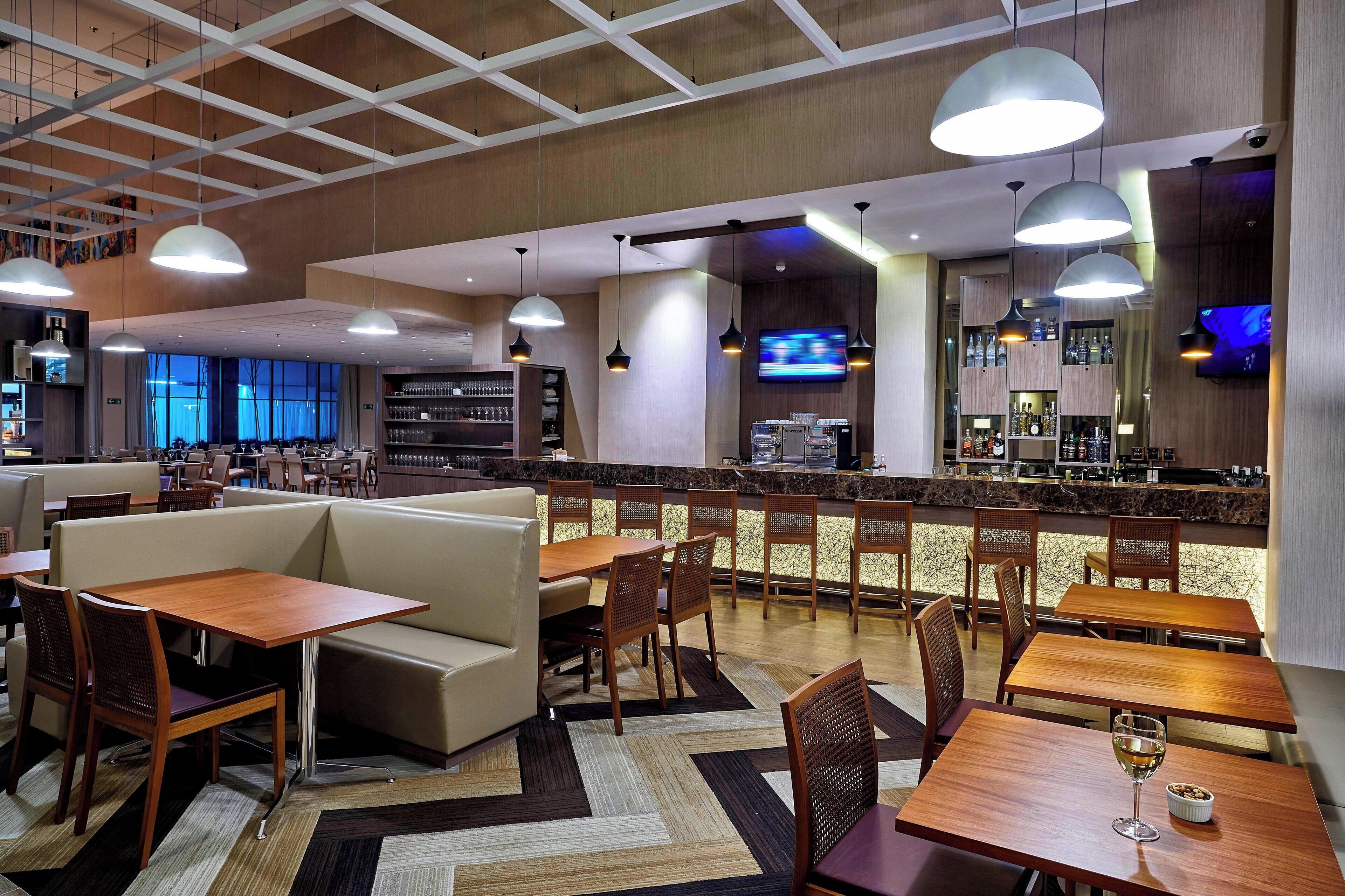 Um hall de hotel moderno com área de bar, espaços para sentar e uma tela de televisão.