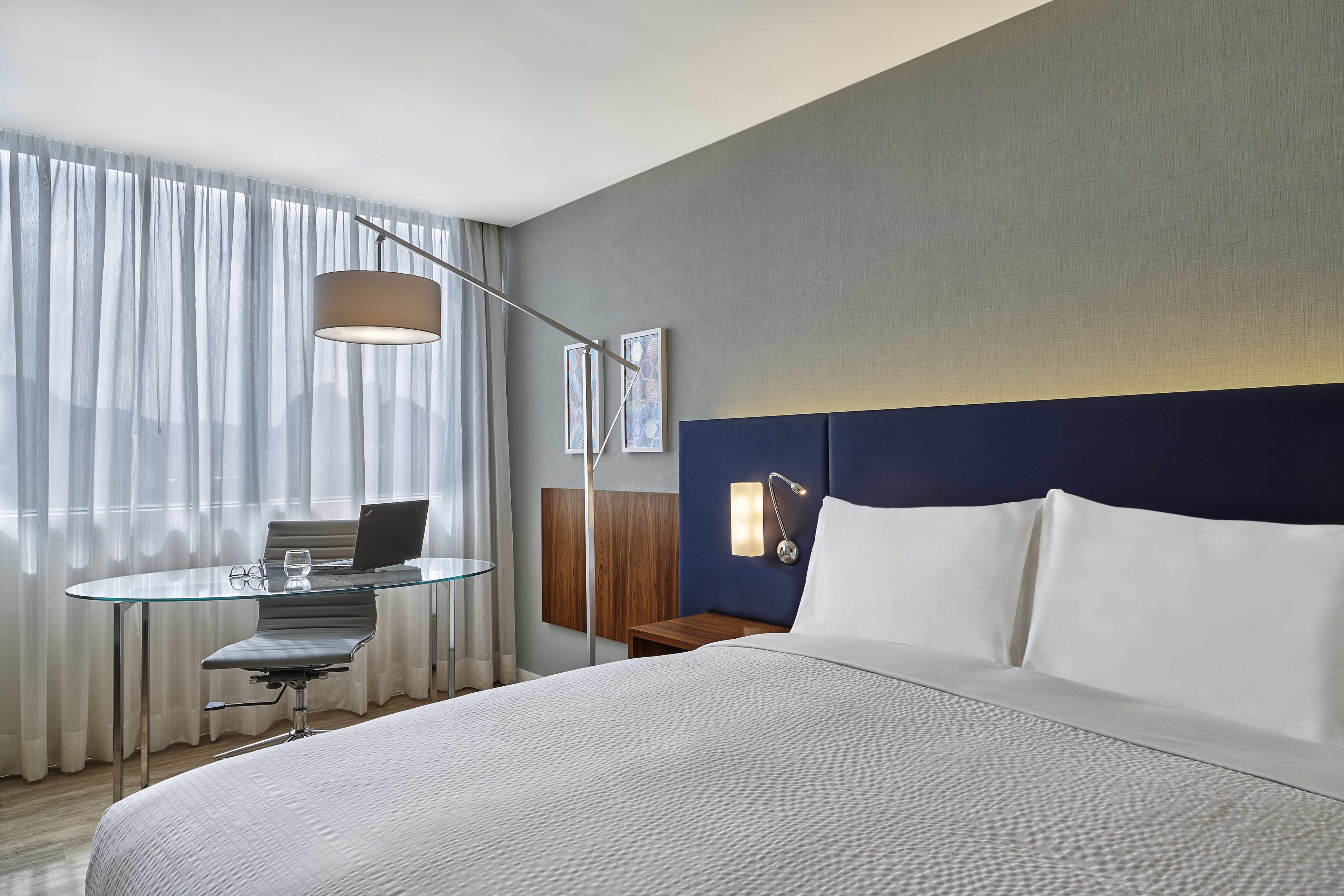A hotel room with a large bed, a glass table with a chair, a desk, and a lamp.