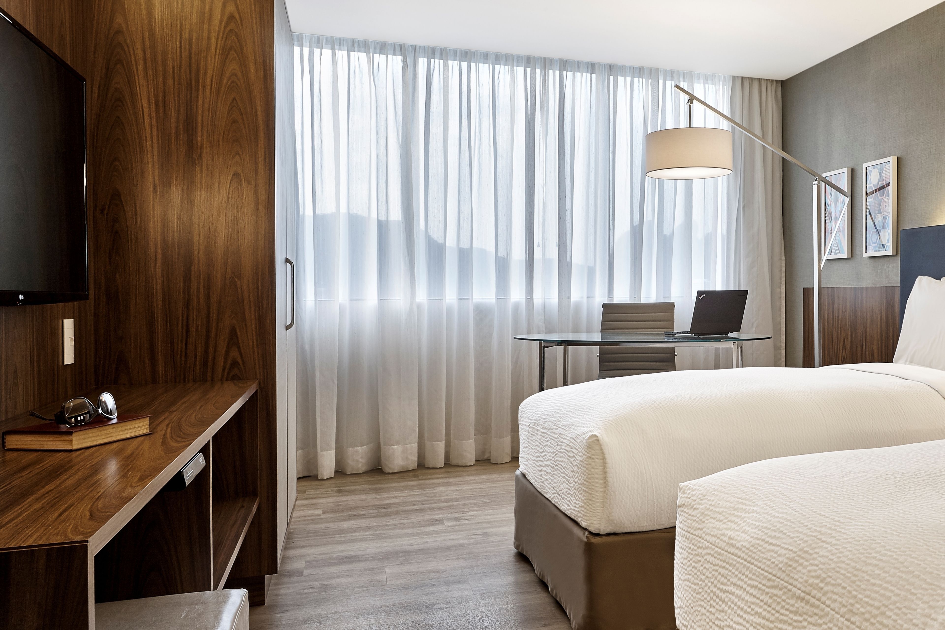 A hotel room with a wooden wall, a large window with sheer curtains, a desk with a laptop, a bed with white bedding, and a floor lamp.