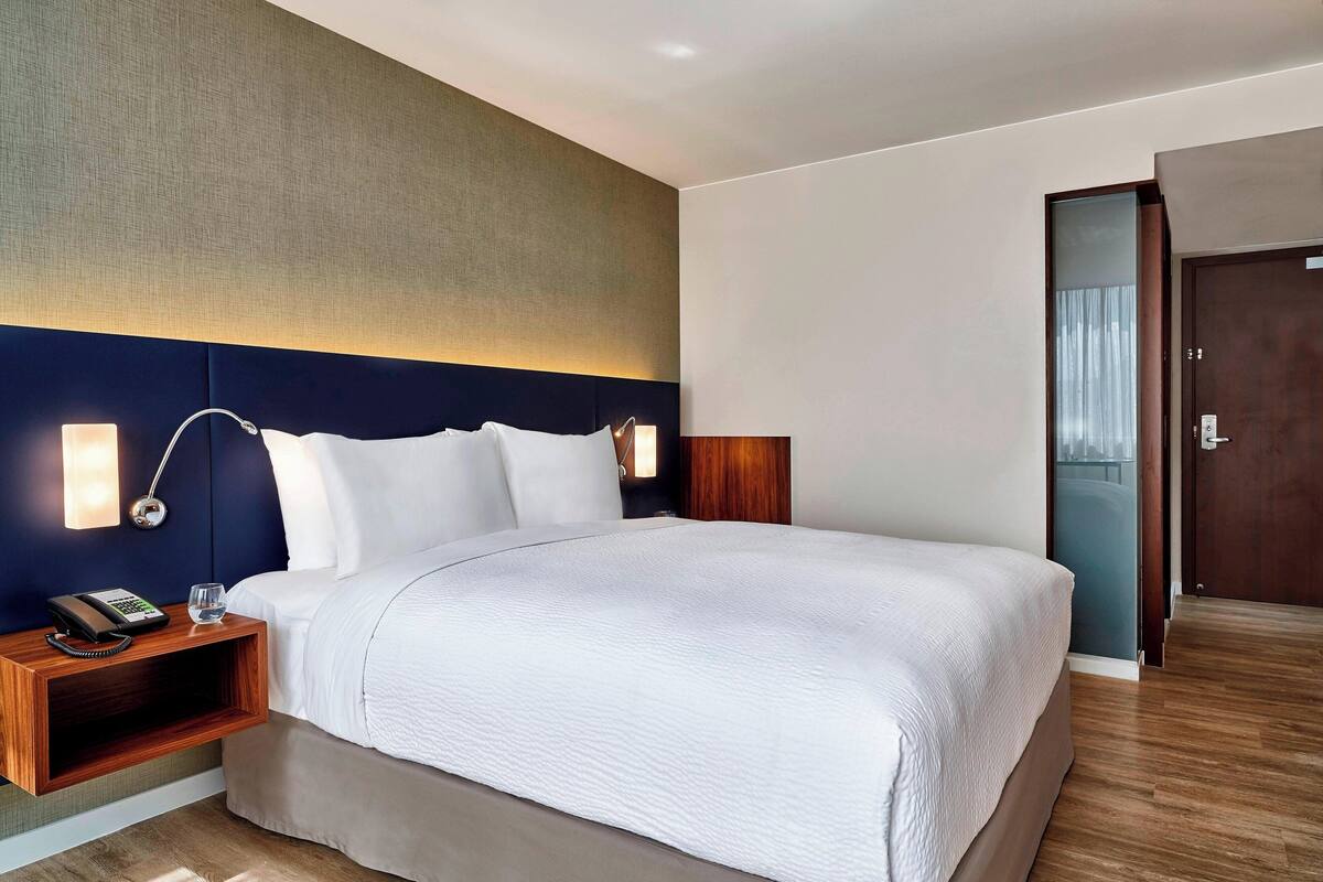 A modern hotel room with a large bed, bedside lamps, a nightstand, and a wooden headboard.