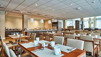 A spacious hotel dining area with multiple tables and chairs, a central buffet, and a modern ceiling design.