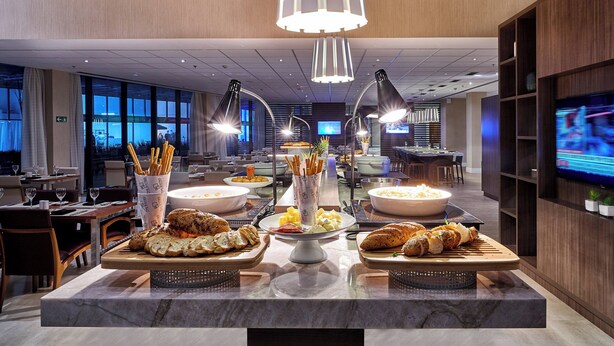 Montaje de bufé con diferentes opciones de comida, incluyendo pan y pastelería, en un comedor moderno de hotel con amplias ventanas y obras de arte en las paredes.