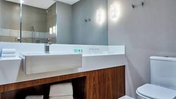 A modern bathroom with a large mirror, white sink, and toilet. There are towels on the counter and wooden shelves below.