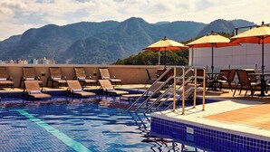 Una piscina en la azotea con sillones y sombrillas, rodeada de montañas.