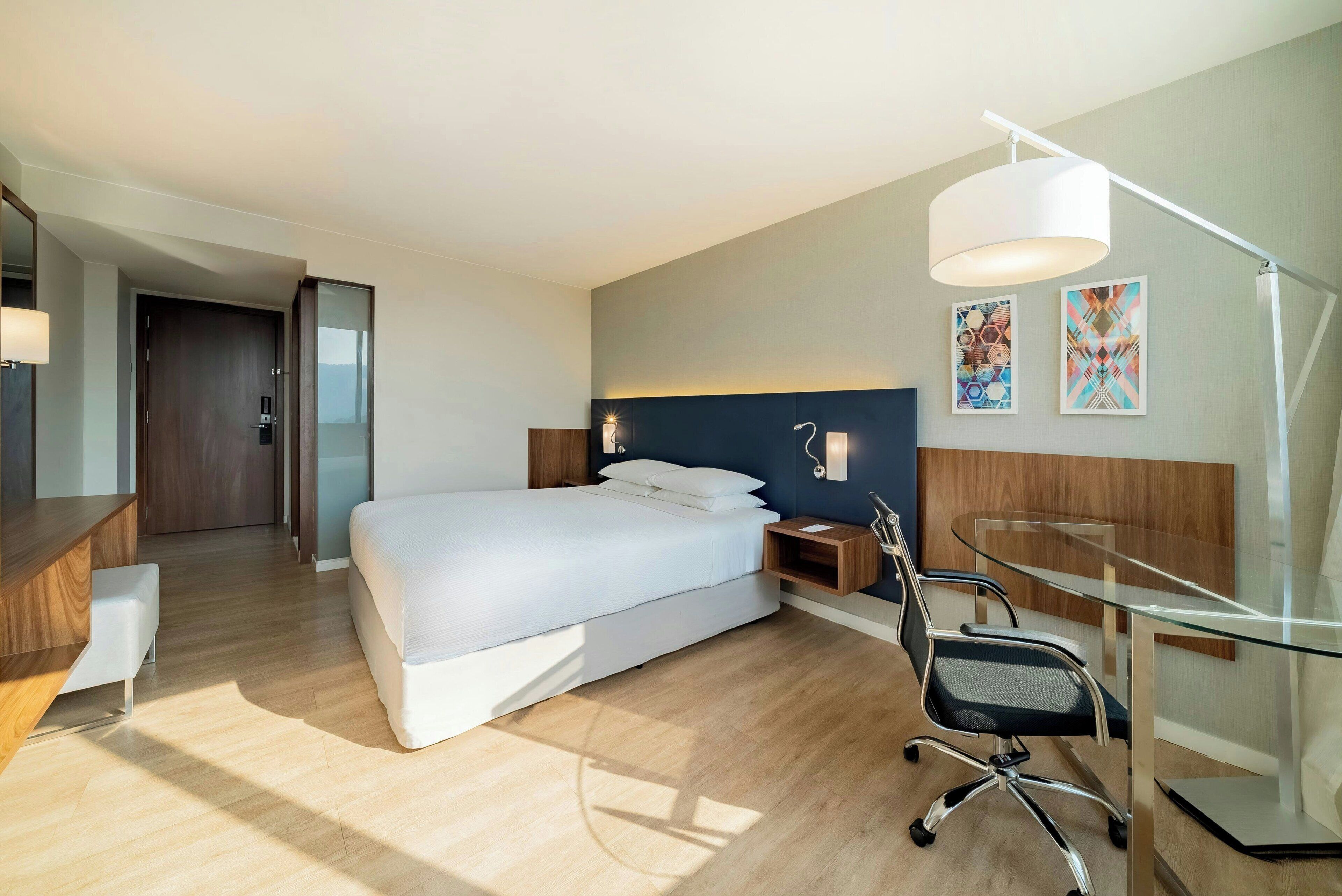 Ett modernt hotellrum med en säng, ett skrivbord, en stol och en skrivbordslampa.